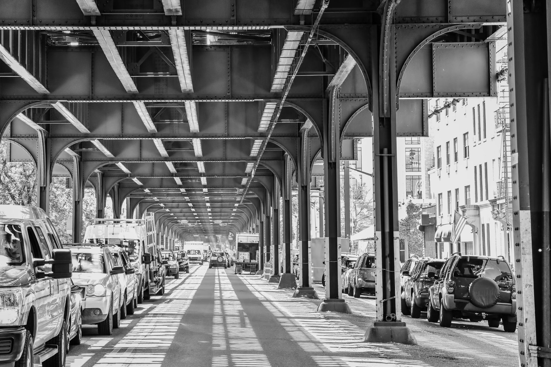 Black-and-white view of a city street under an elevated train track, lined with parked cars.