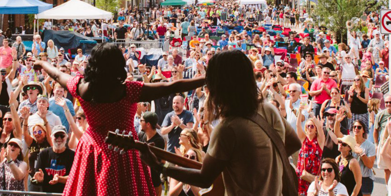 Rhythm & Roots Reunion Festival Website A woman in a red dress and a man with long hair are playing guitars and singing in front of a large crowd at the Rhythm & Roots Reunion Festival Stage in Bristol