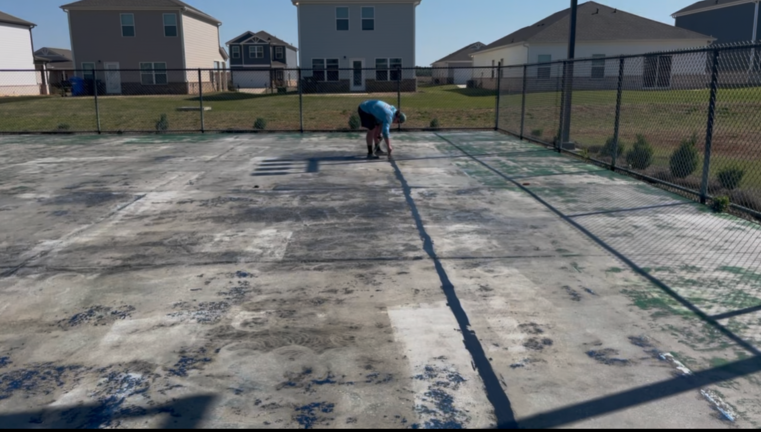 A person bends down on a weathered, light-colored concrete court near a chain-link fence in a residential neighborhood.