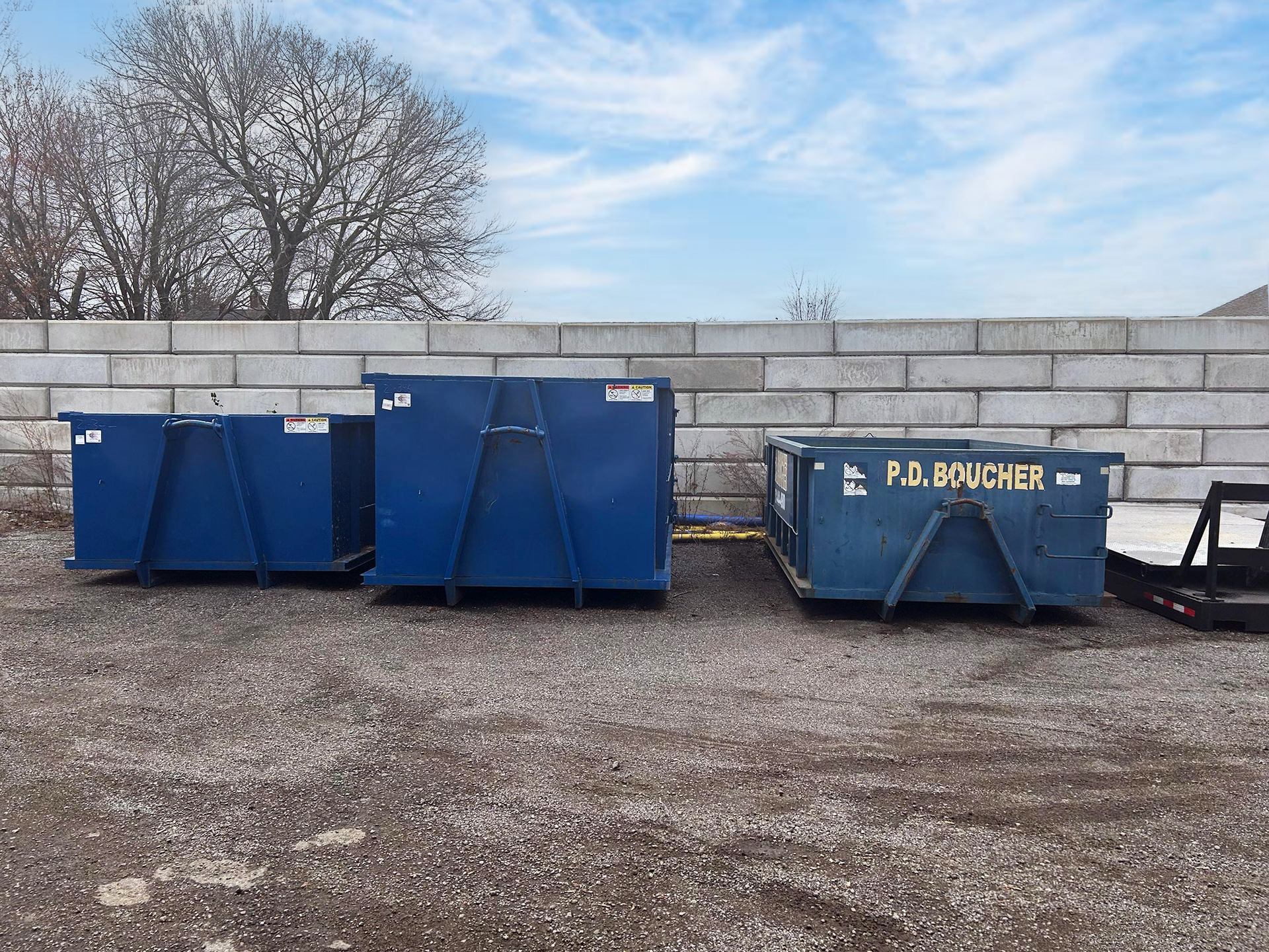 Three blue dumpsters of varying sizes sit on gravel in front of a concrete wall.