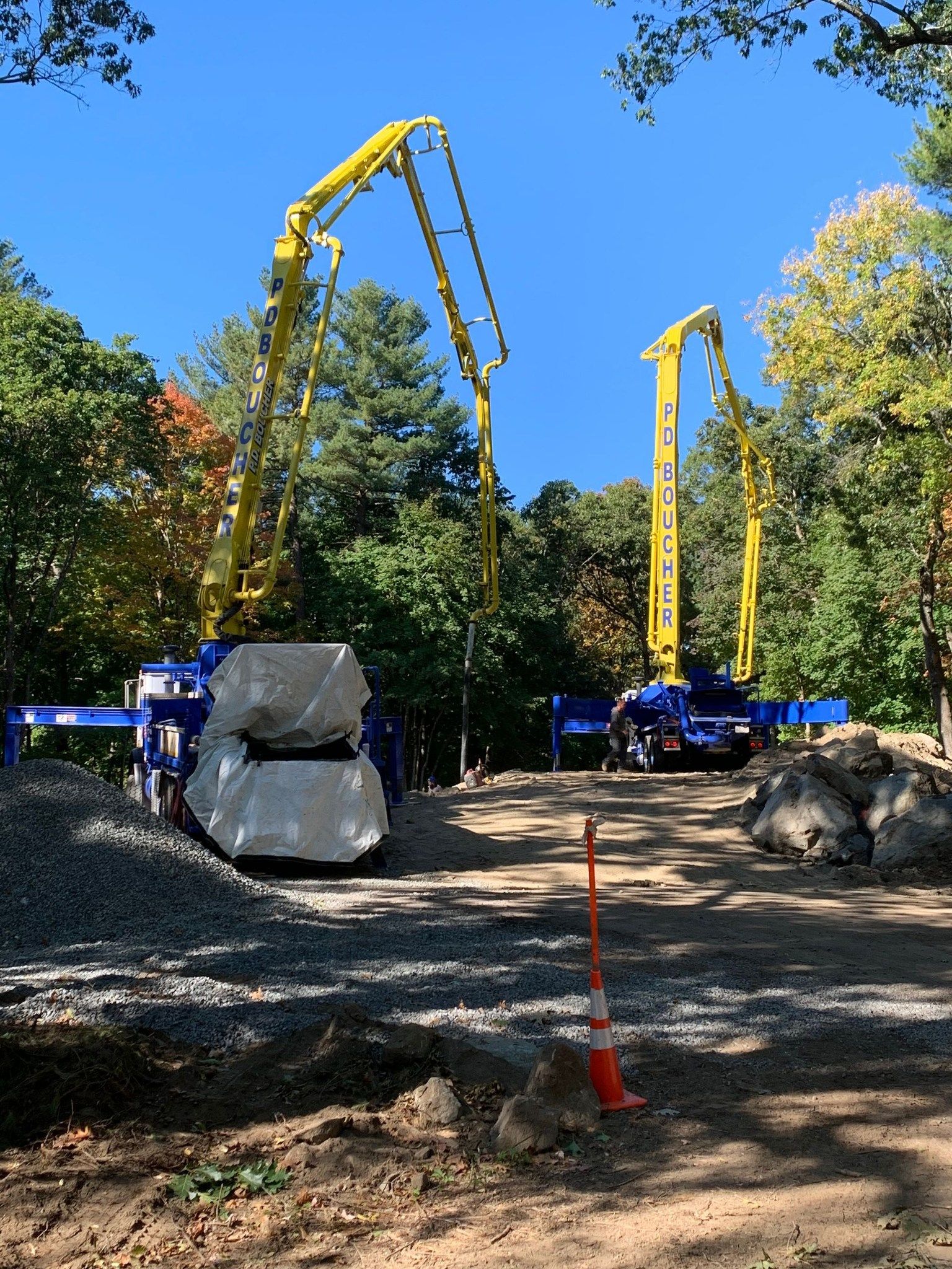 Two concrete pump trucks with raised yellow booms on a construction site. Blue sky and trees in the background.