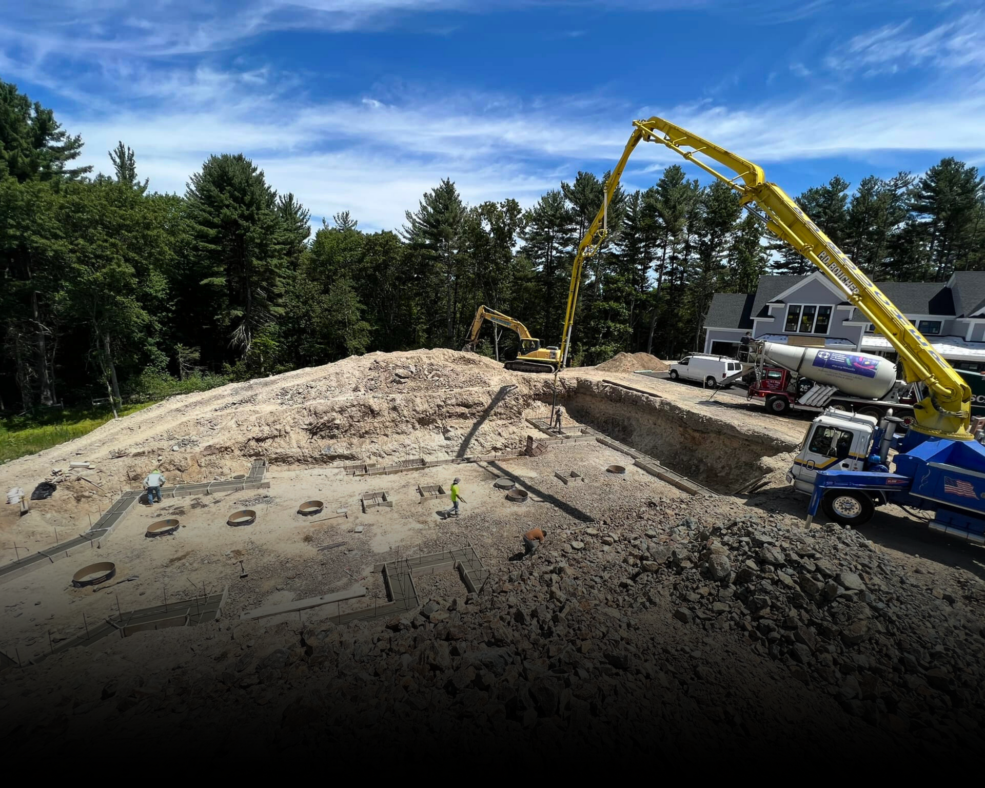 Construction site with concrete pump truck pouring concrete into foundation. Yellow excavator and trees in the background.