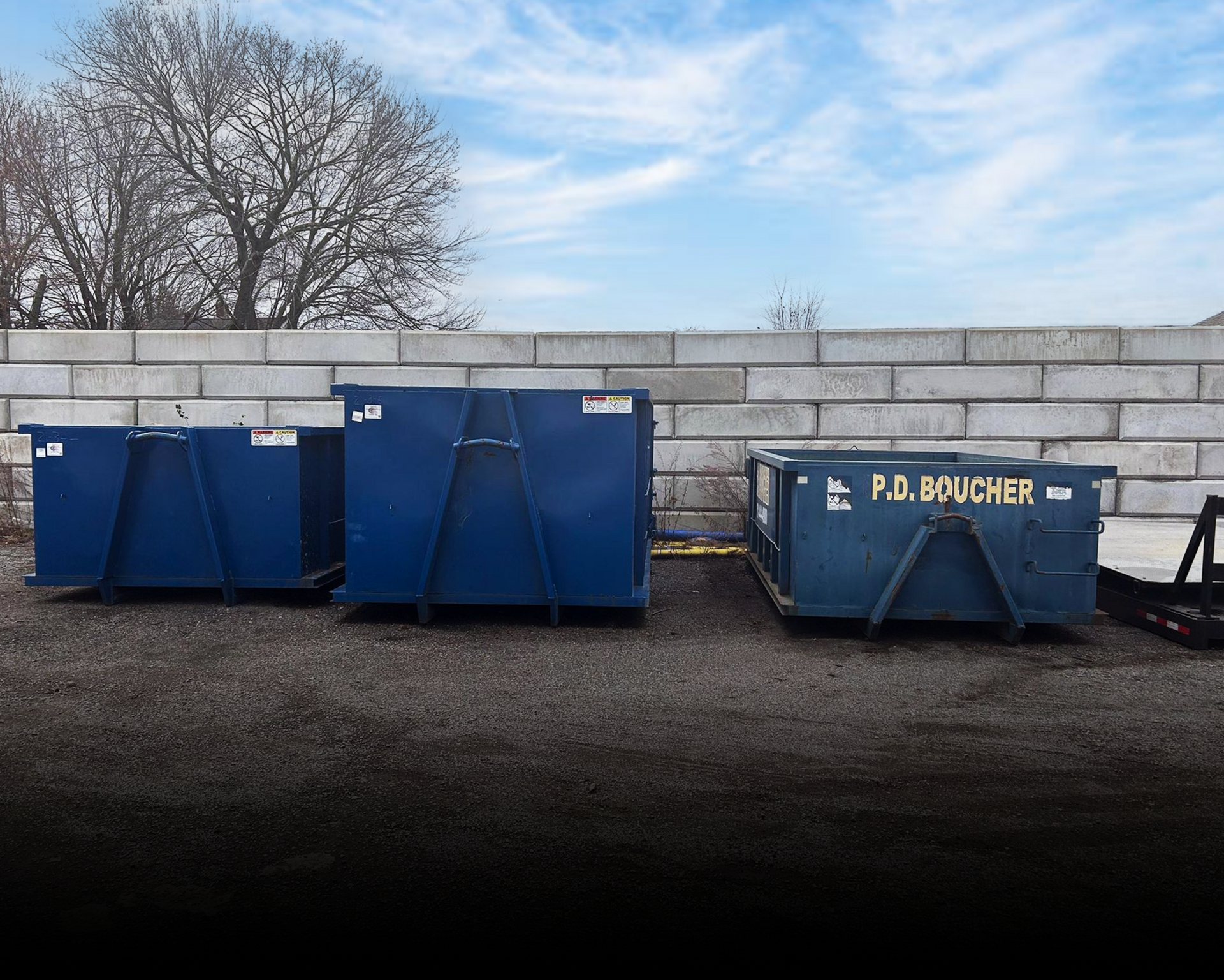Three blue dumpsters in a gravel lot next to a concrete wall. The sky is partly cloudy.