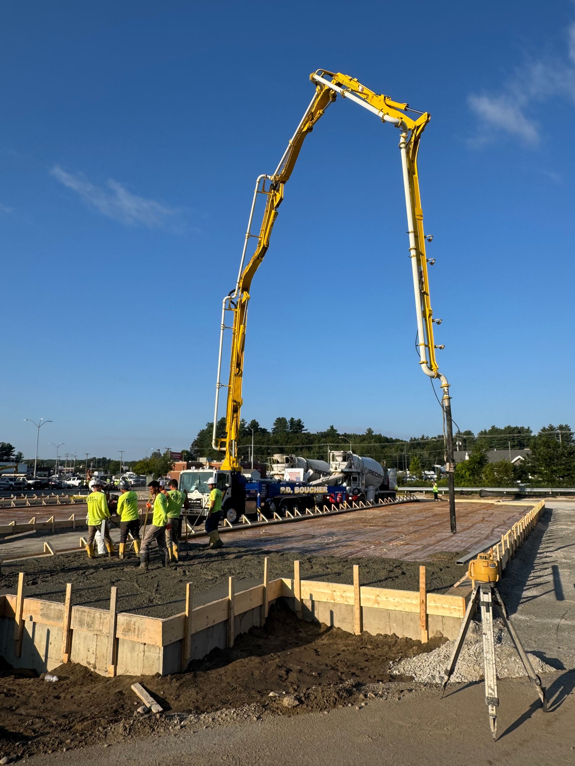 Yellow concrete pump pours concrete into a framed area. Workers in yellow vests stand nearby. Clear blue sky.