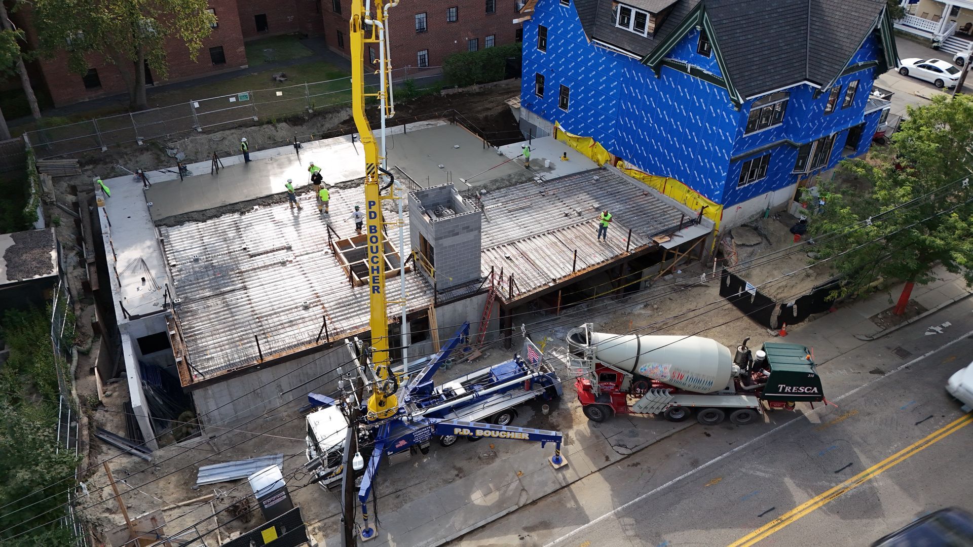 Construction site with concrete pump truck pouring concrete into a foundation. Blue house nearby.