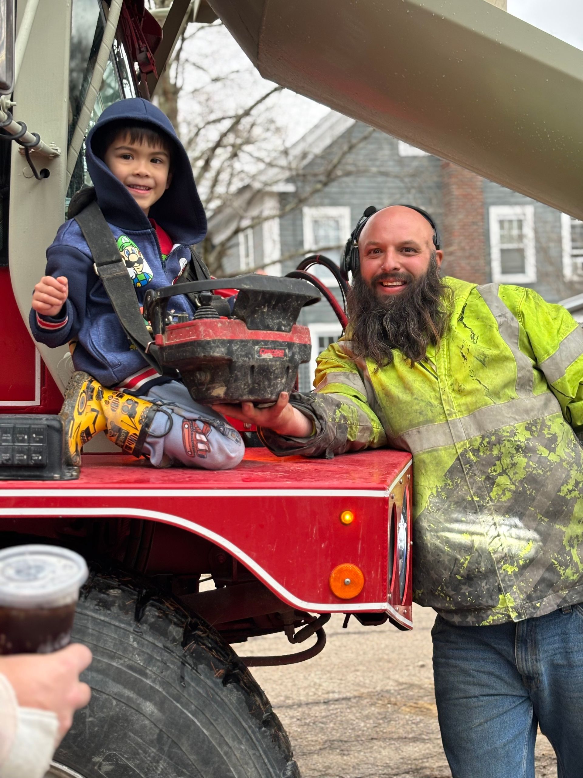 Boy in hoodie sits on truck with man in safety vest. Both smiling; boy holds something.