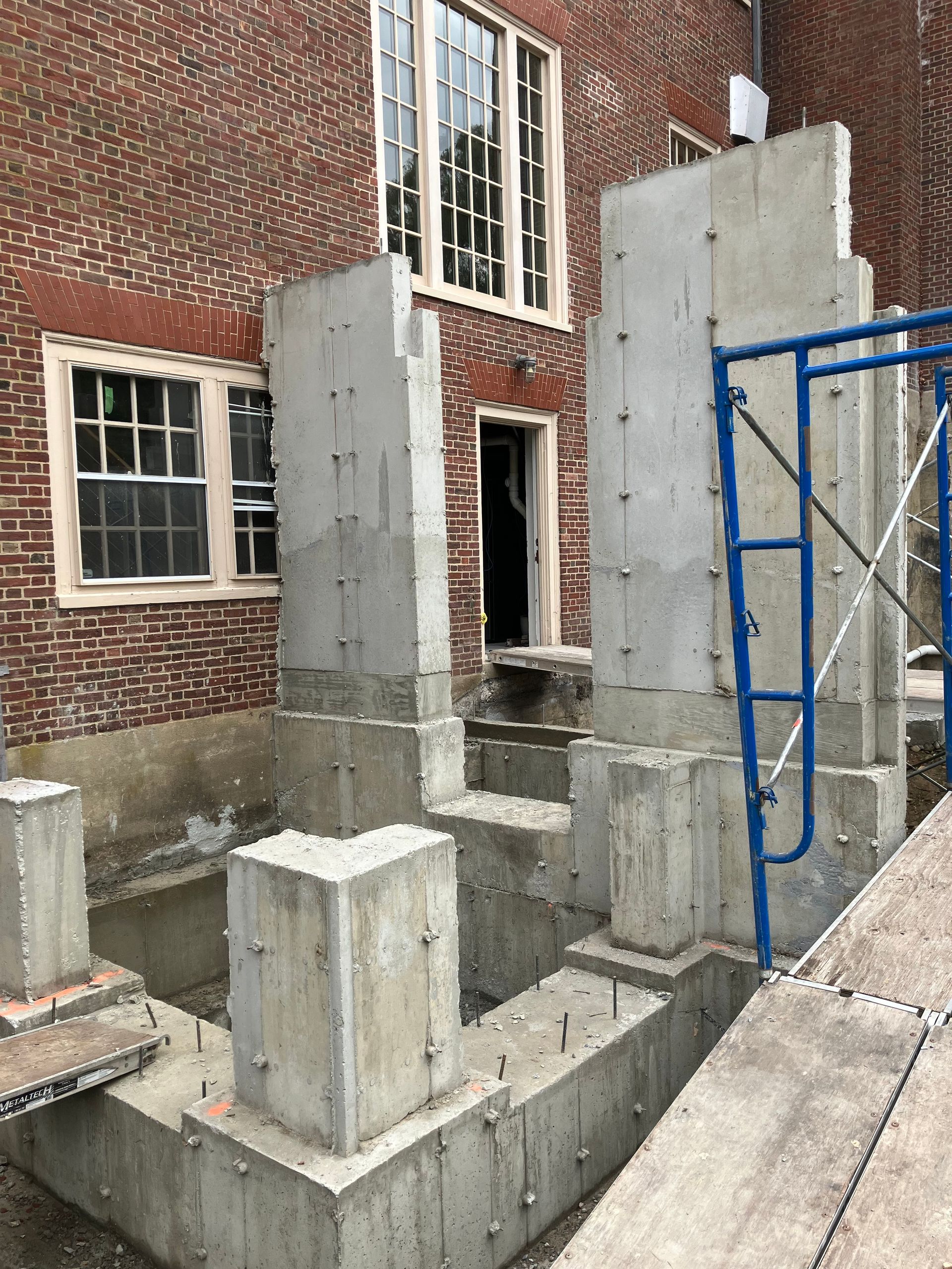 Concrete foundation and pillars under construction against a brick building with windows and a doorway.