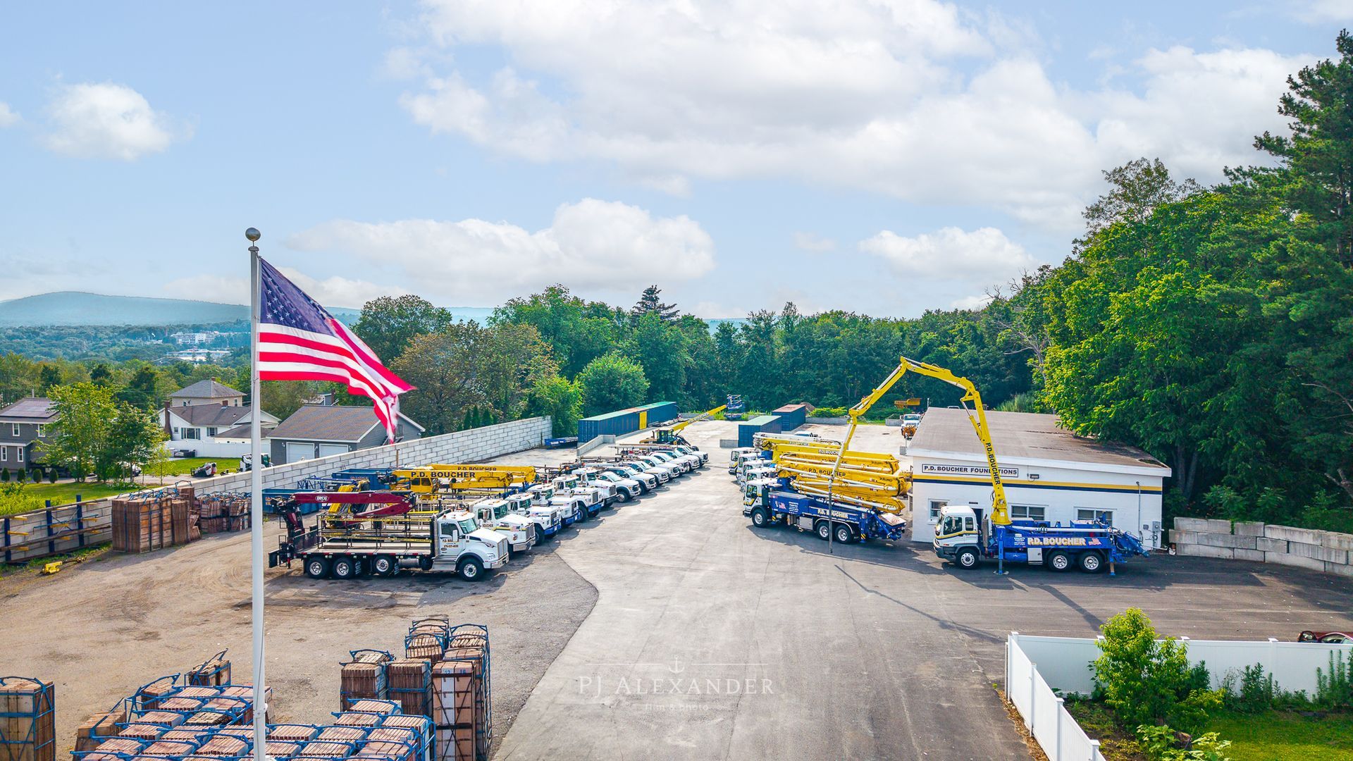 Fleet of work trucks parked outside a building, American flag in foreground, mountain range in the distance, sunny day.