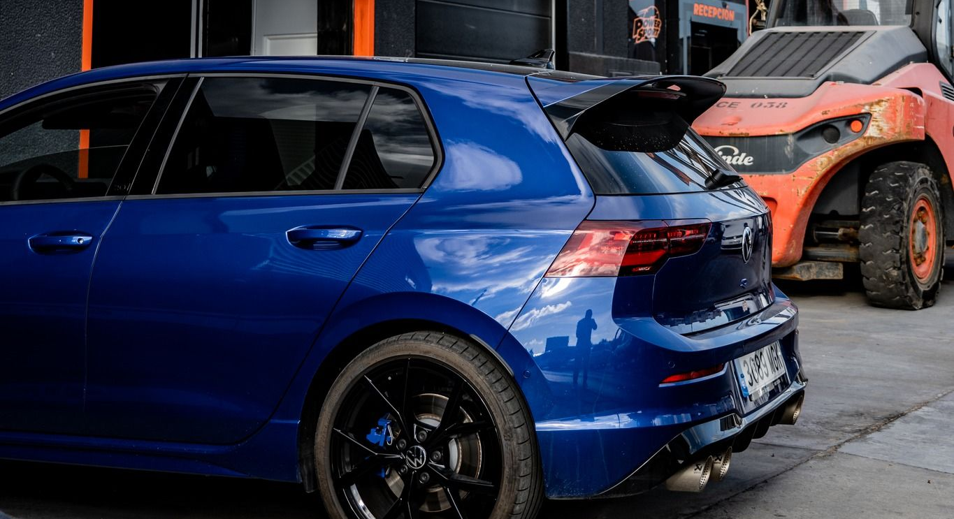 Un Volkswagen Golf hatchback azul con alerón negro estacionado junto a un vehículo de construcción naranja.
