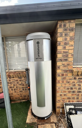 White cylindrical water tank mounted beside a brick house under a roof overhang.
