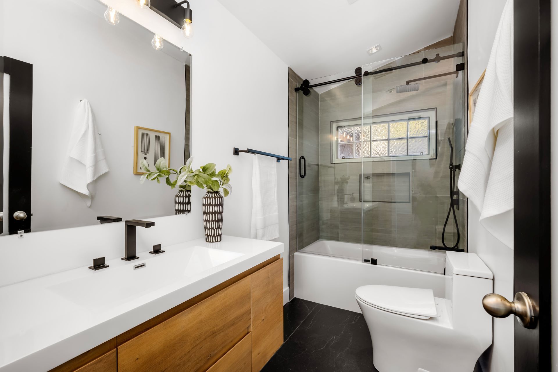 Modern bathroom with white vanity, wood cabinet, glass shower, black fixtures, and a white toilet.