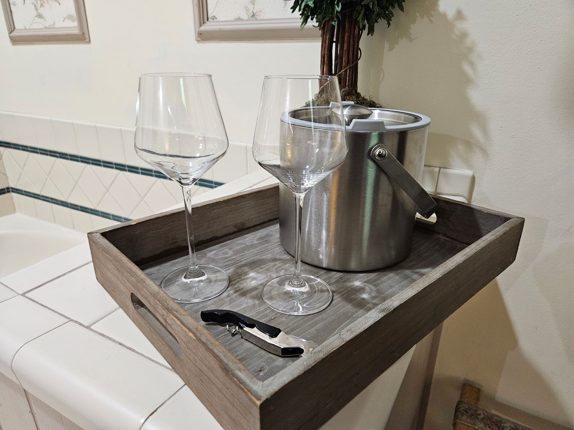 A wooden tray with wine glasses, an ice bucket, and a corkscrew on a countertop.