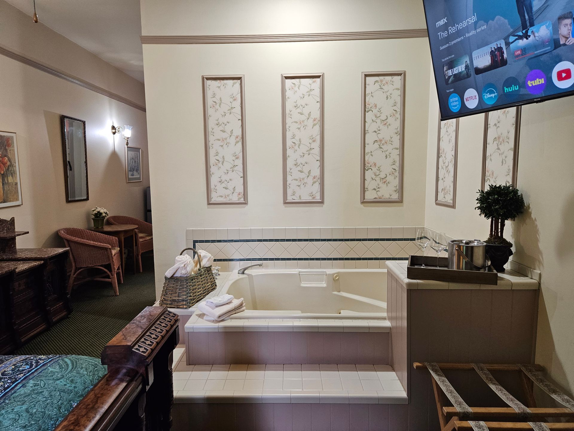 Hotel room with a jacuzzi, TV, art, seating area, and luggage rack. Beige and off-white tones.