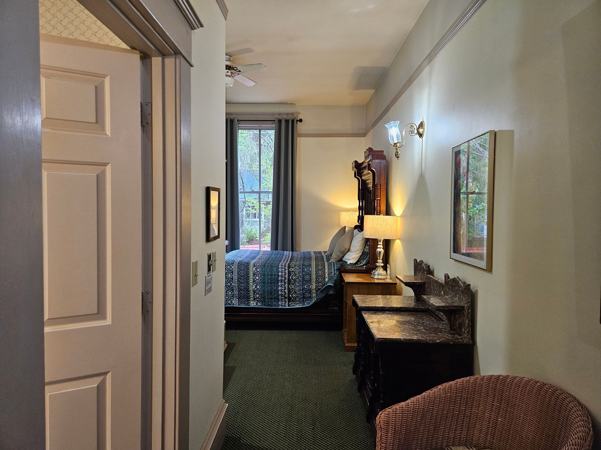 Hallway with view of bedroom: bed, nightstands, chair, green carpet, white door.