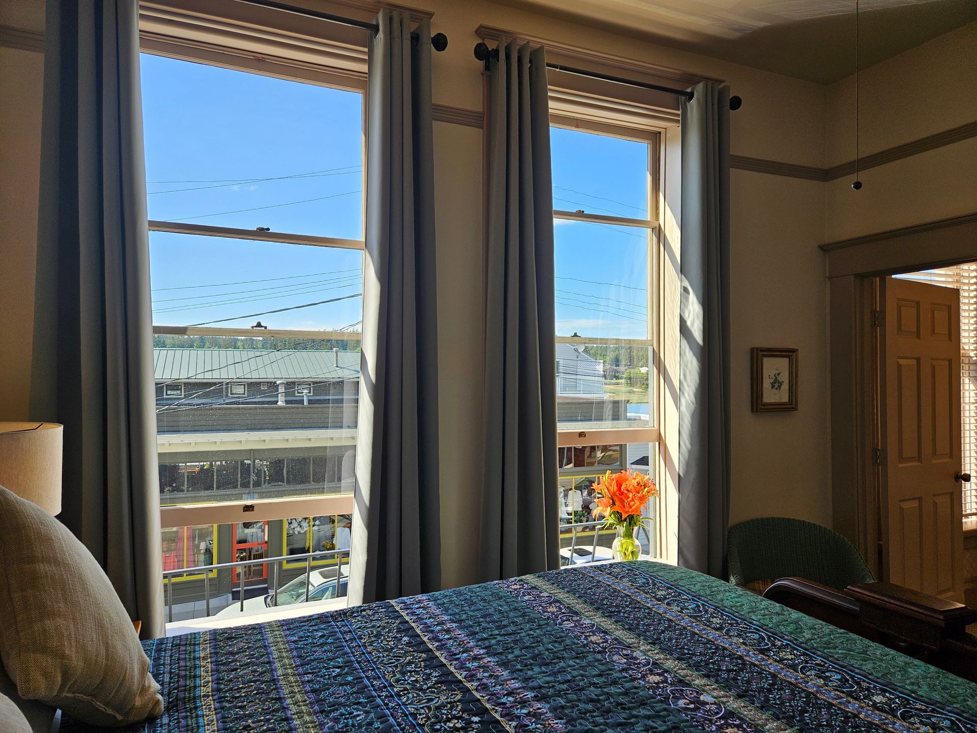 Bedroom with large windows overlooking a city street. Light blue curtains frame the view.