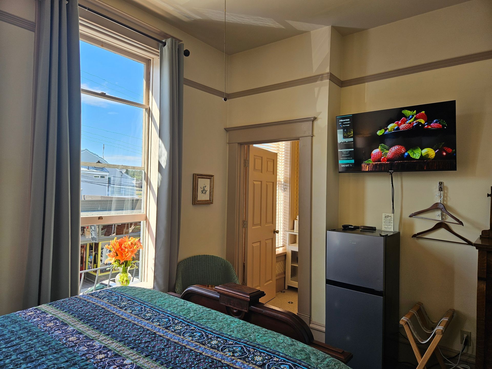 Bedroom with open window, bed, TV, mini-fridge, and doorway to another room.