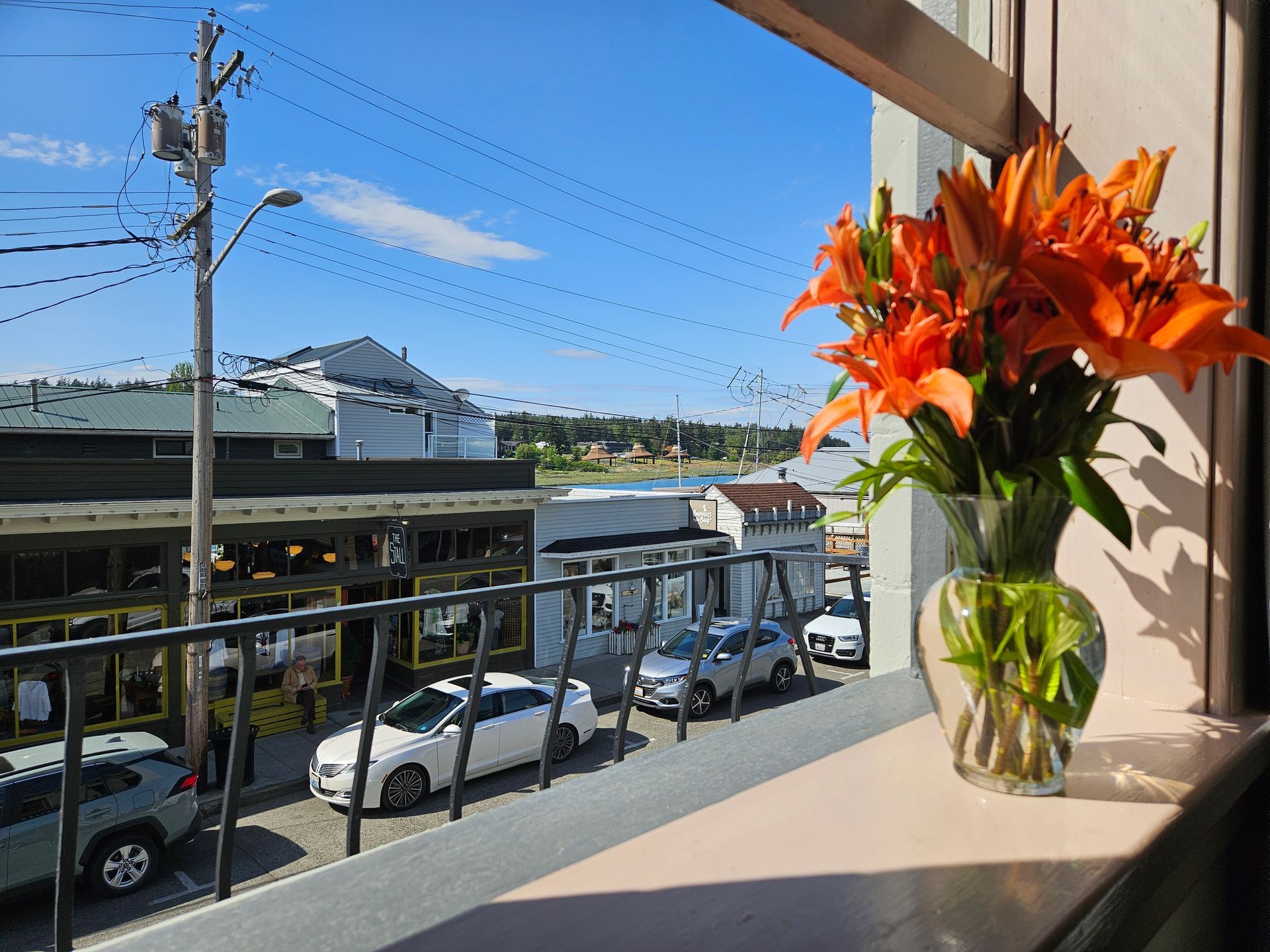 Orange lilies in a vase on a windowsill overlook a sunny street with parked cars and buildings, blue sky.