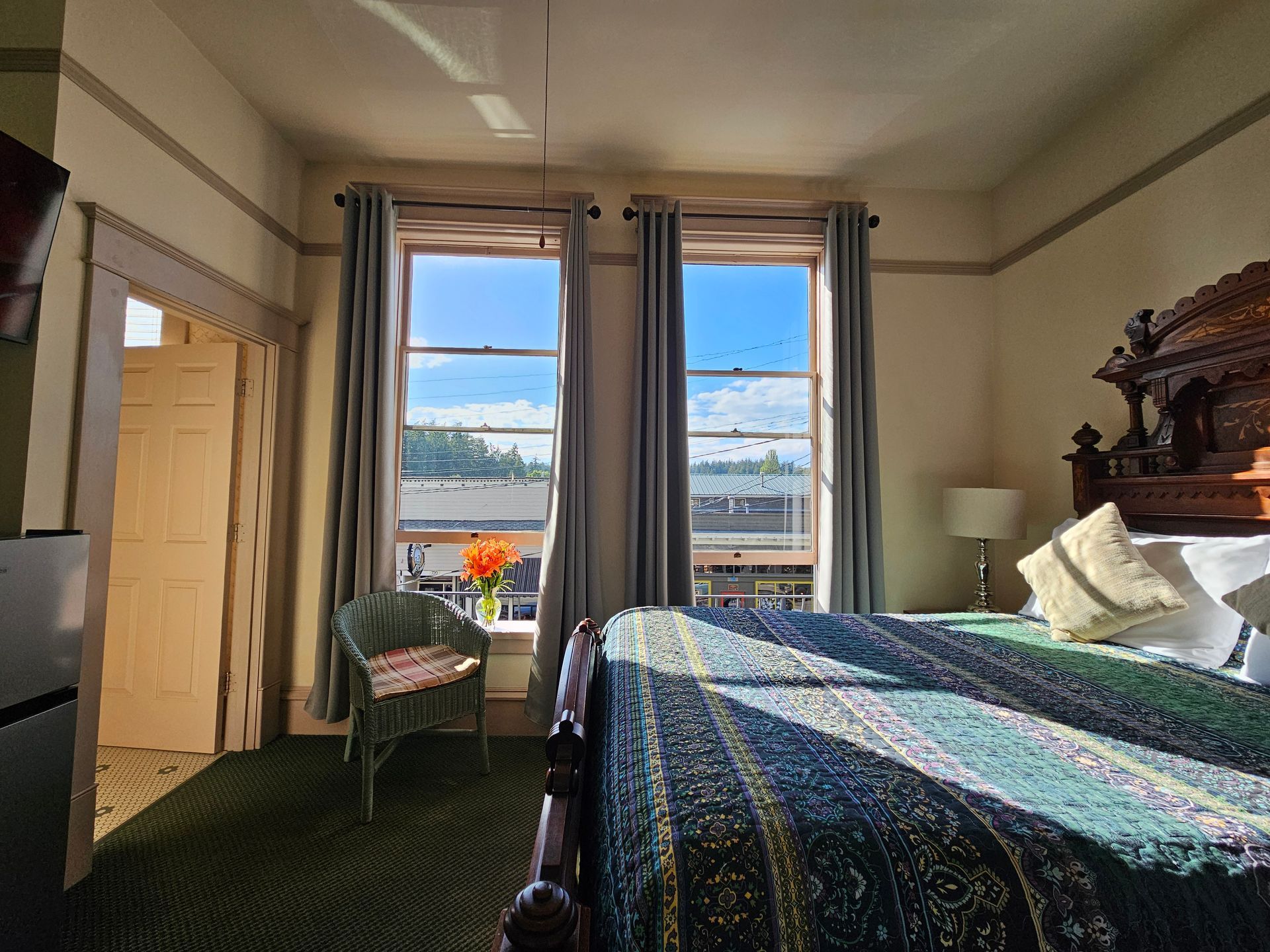 Bedroom with large bed, windows, small chair, and open doorway; sunlight streams in.