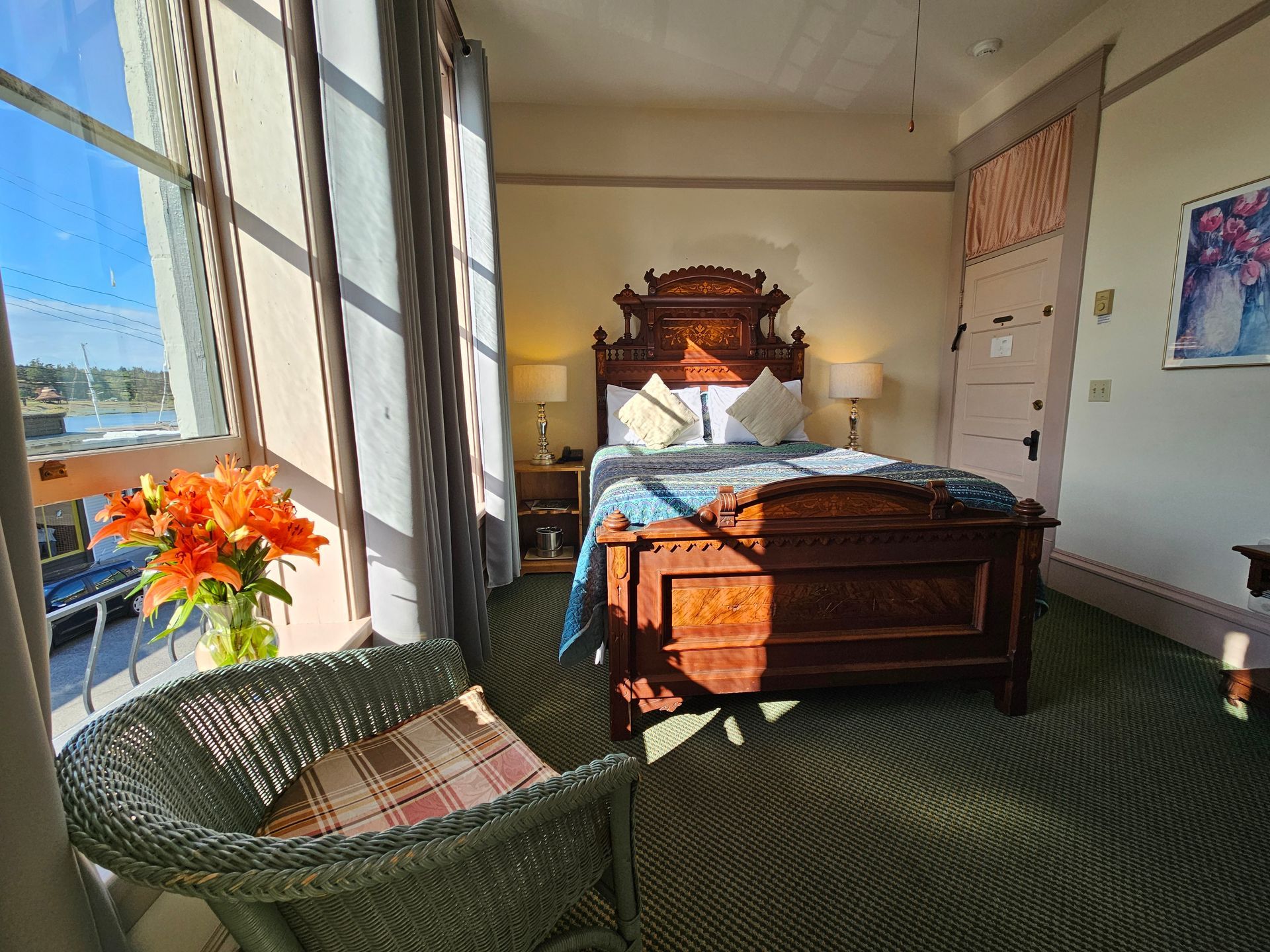 Bedroom with ornate wooden bed, wicker chair by window, and sunlight. Orange flowers on windowsill.