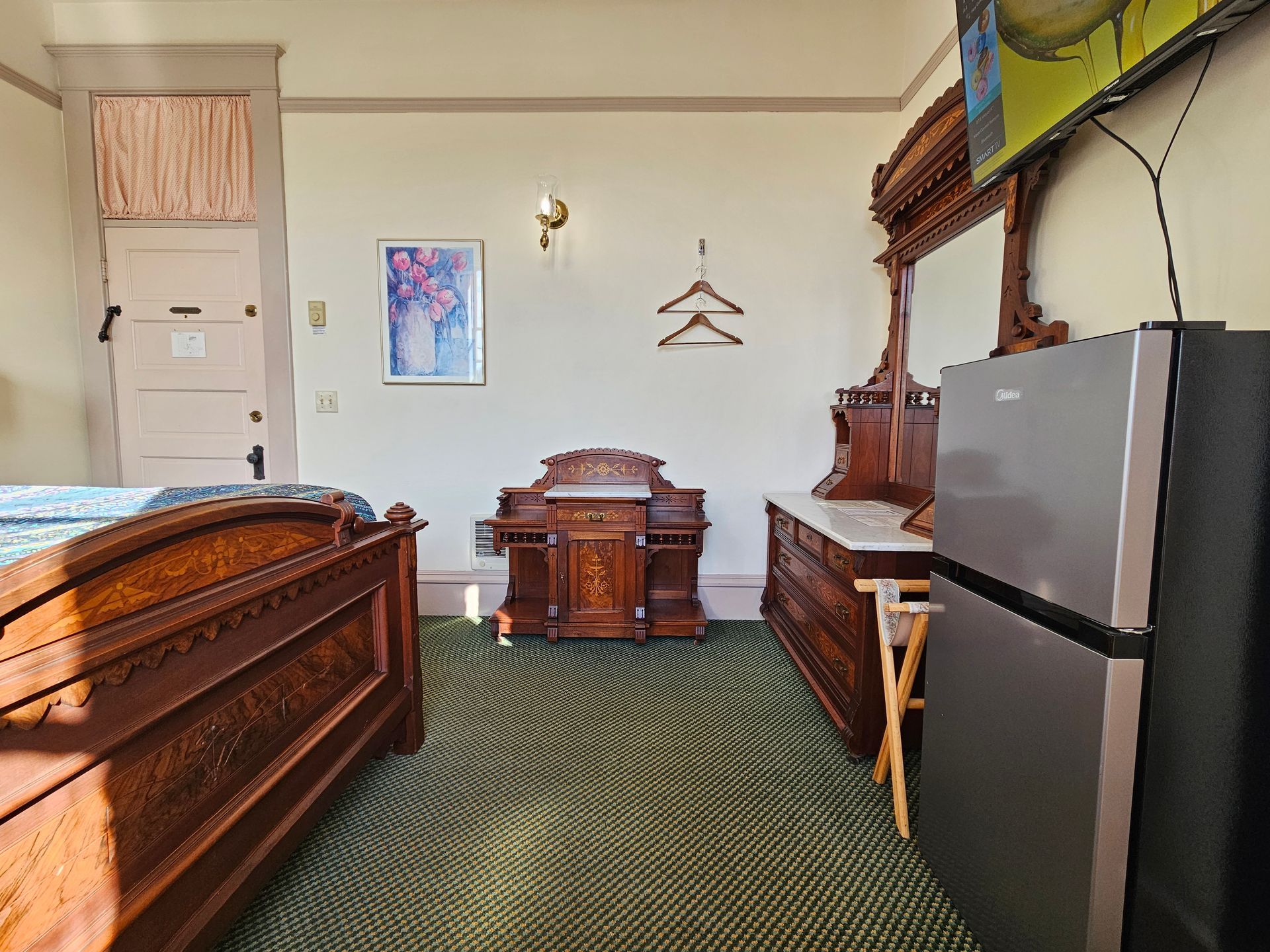 Bedroom with antique furniture: bed, dresser, nightstand, and refrigerator. Green patterned carpet, light walls.