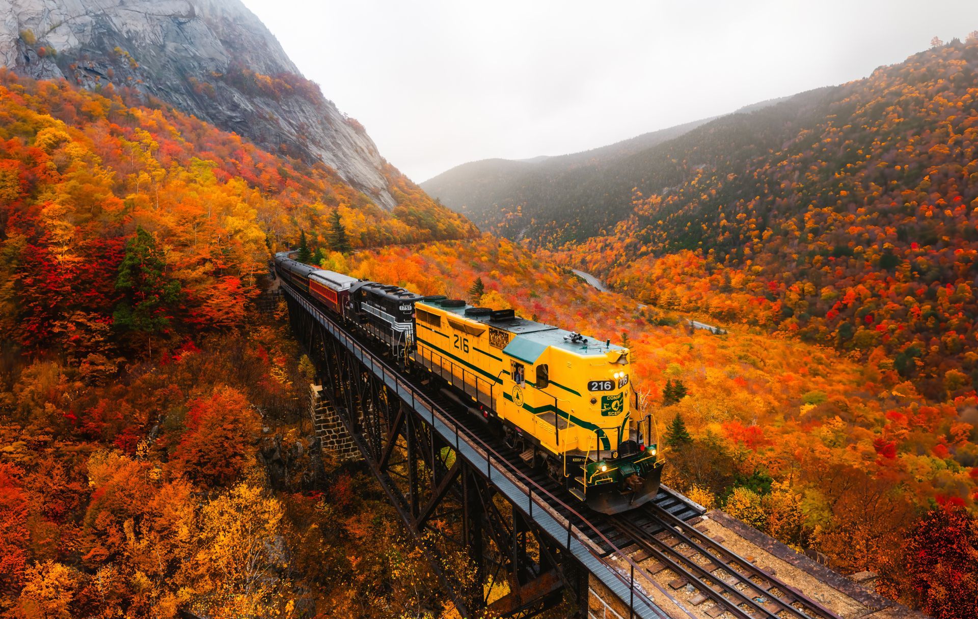 A yellow train crossing a bridge, surrounded by autumn foliage and mountains.