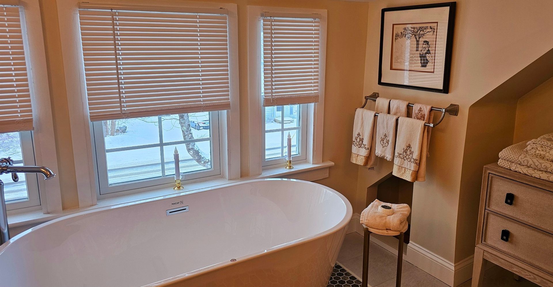 Bathroom with a freestanding white tub under windows with blinds. Towel rack and art on the wall.