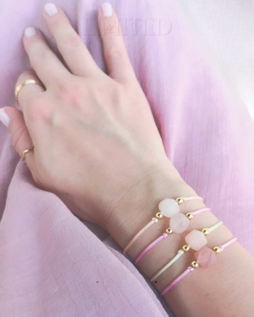 Woman's wrist with three delicate rose quartz bracelets against a pink background.