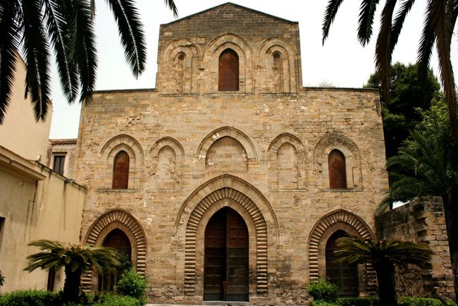 Facciata in pietra della chiesa con porte e finestre ad arco, incorniciata da palme e vegetazione.