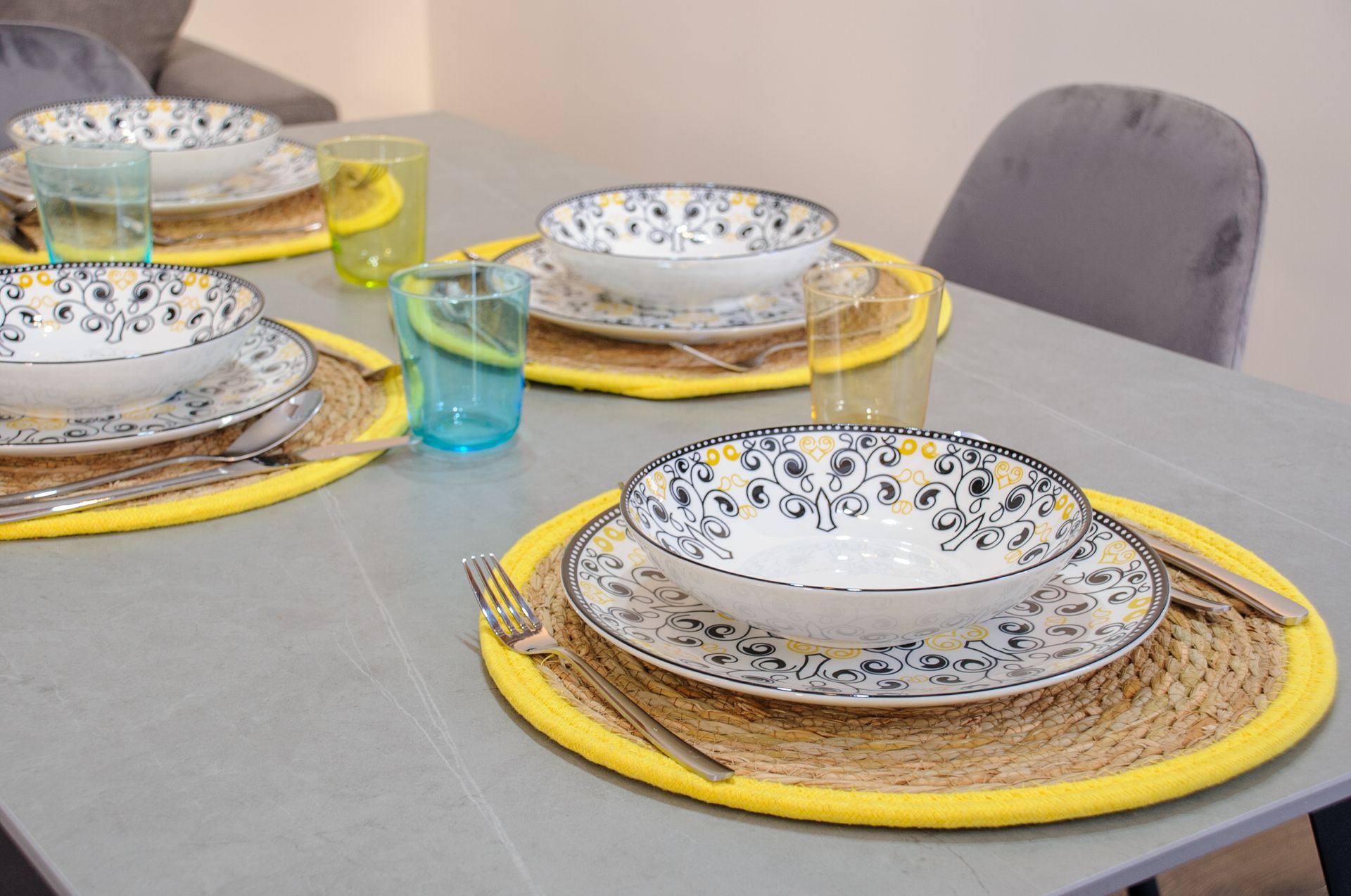 Apparecchia la tavola con tovagliette gialle, piatti decorati e bicchieri blu su una tovaglia grigia.