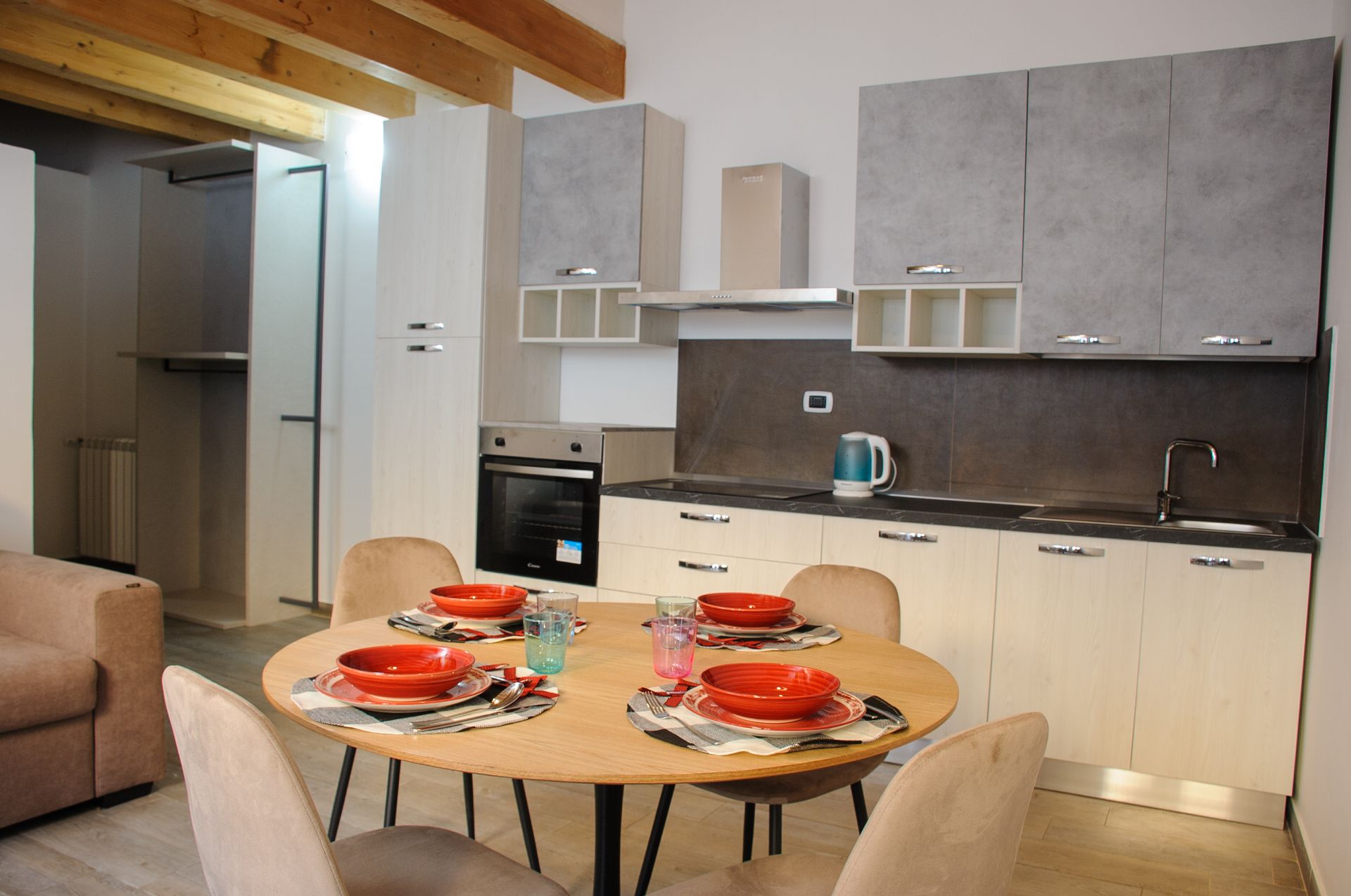 Cucina e zona pranzo moderne con tavolo rotondo per quattro persone, mobili grigi e travi in legno a vista.