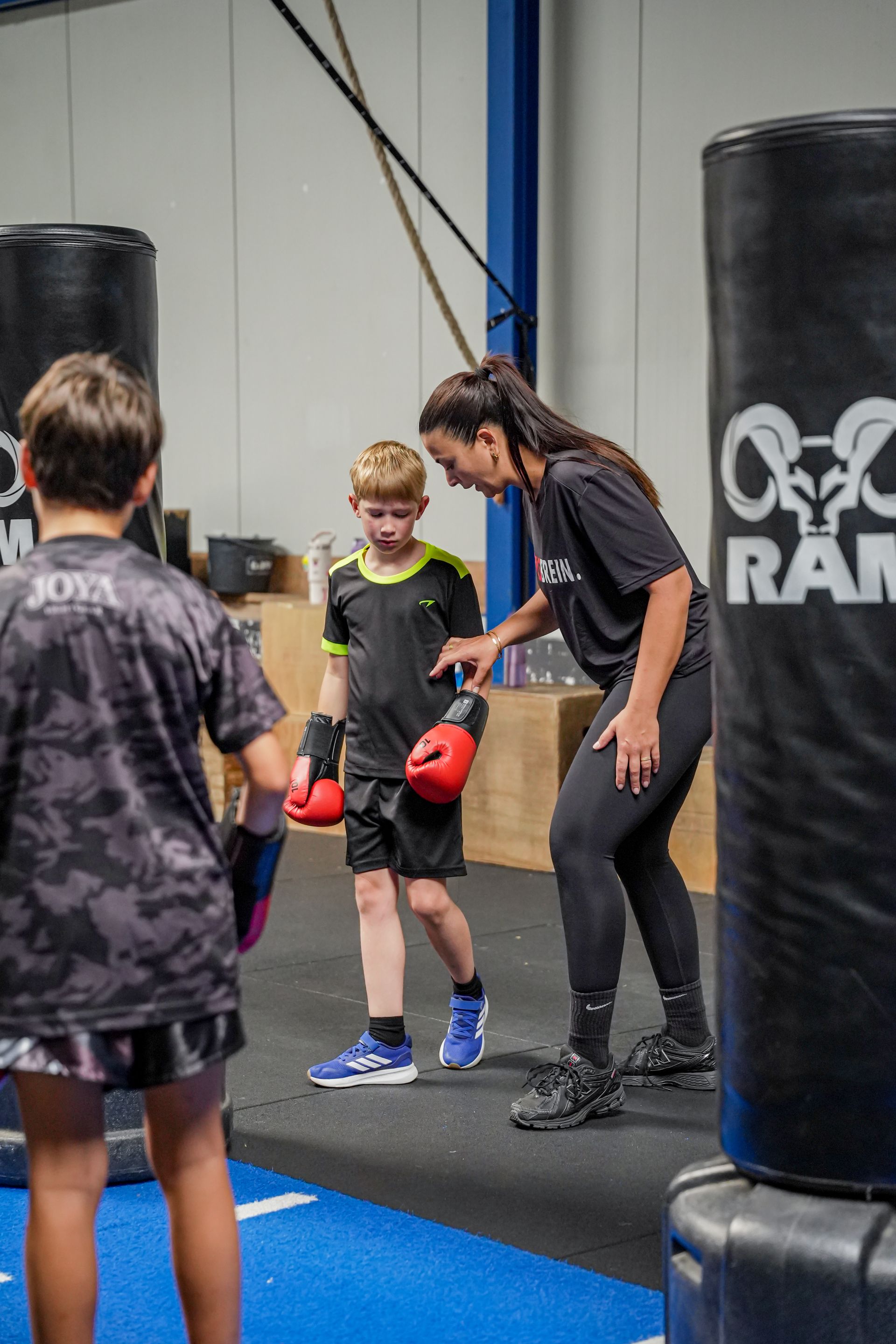 Een persoon die een kind begeleidt bij bokstraining, waarbij het kind op een zwarte, zware bokszak slaat in een sportschool.