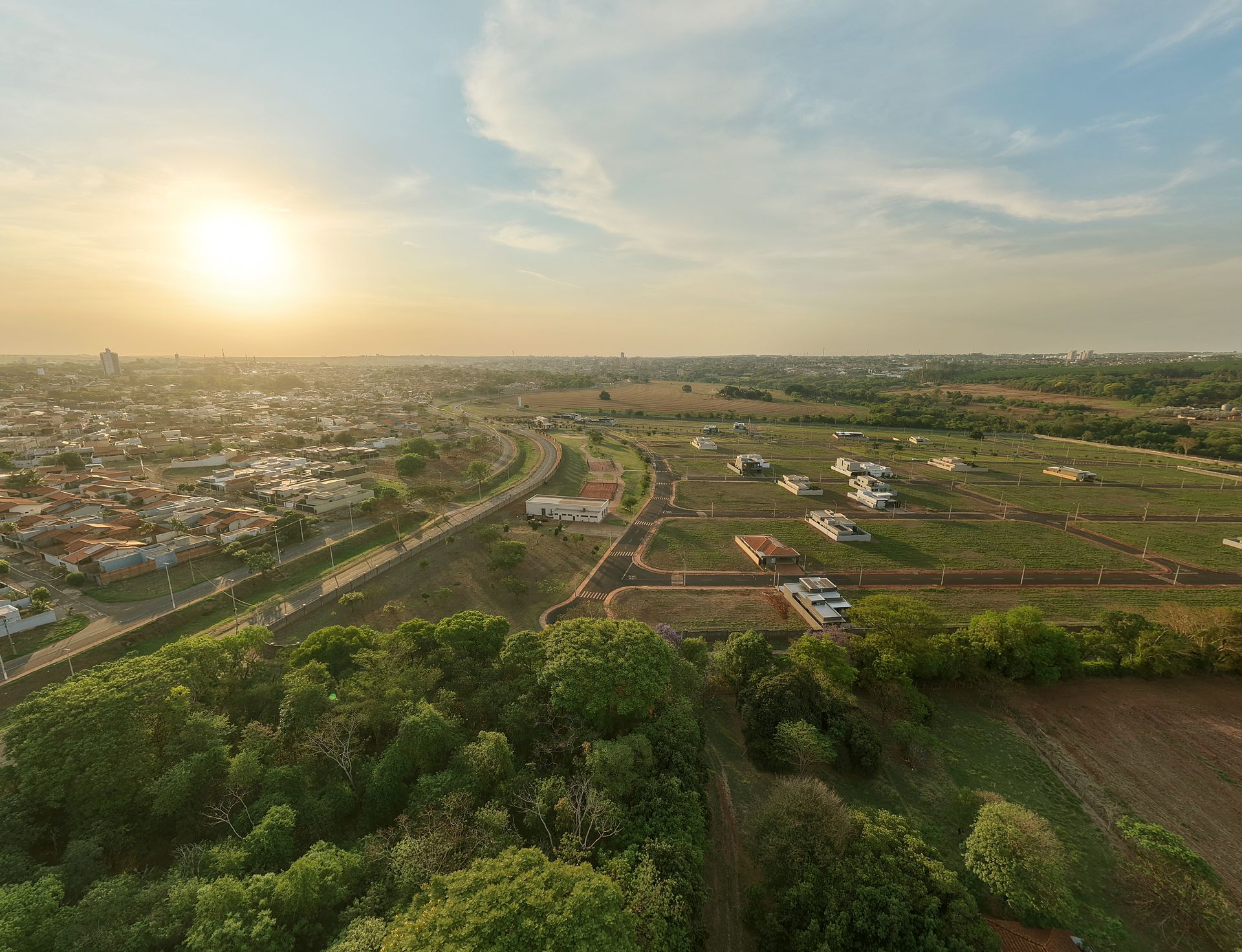 Pôr do sol sobre uma área urbana e rural, com árvores em primeiro plano.