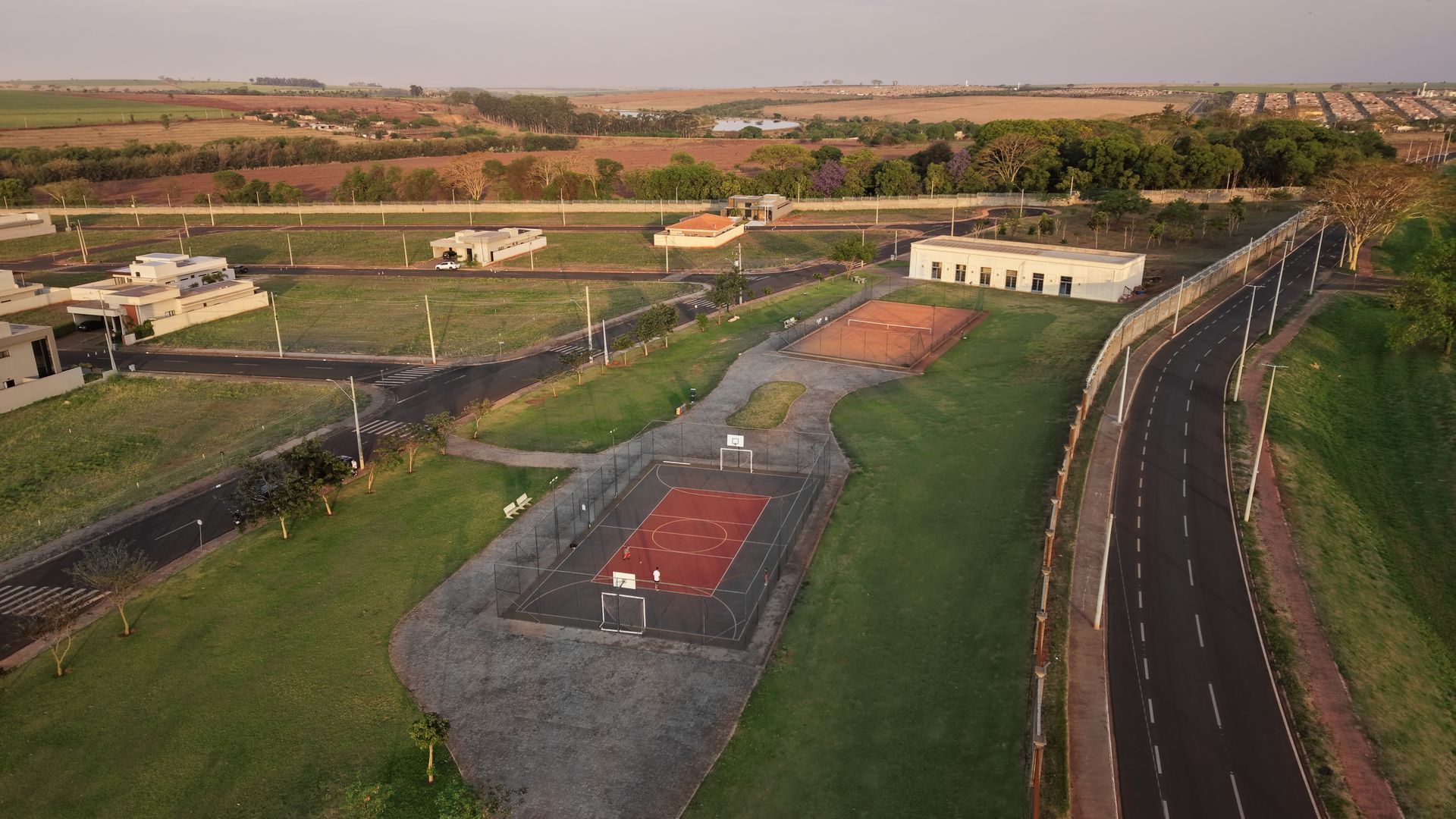 Vista aérea de uma área residencial com quadras esportivas, ruas e casas em uma paisagem gramada sob um céu claro.