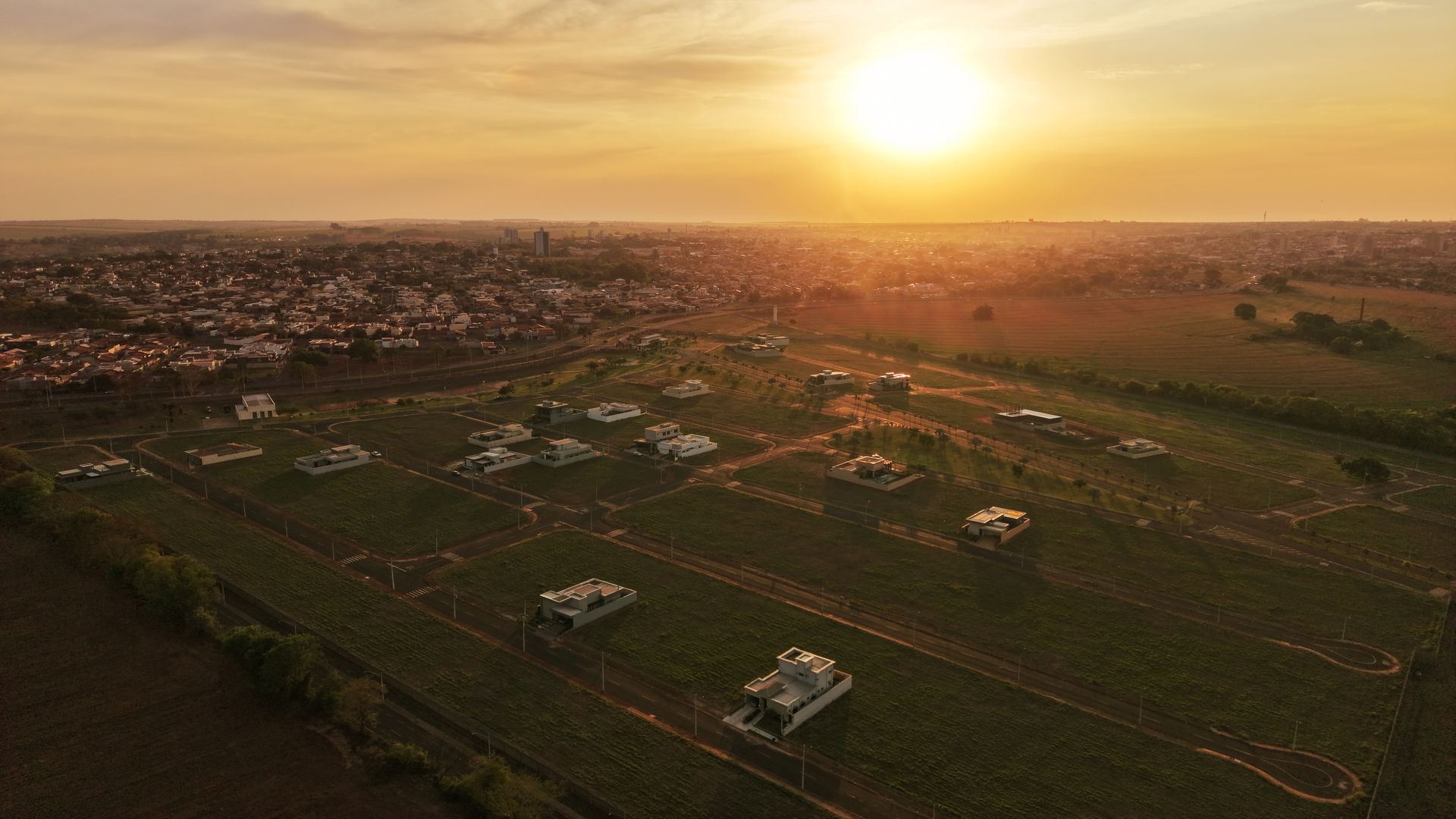 Pôr do sol sobre um campo com casas, estradas e uma cidade ao longe.