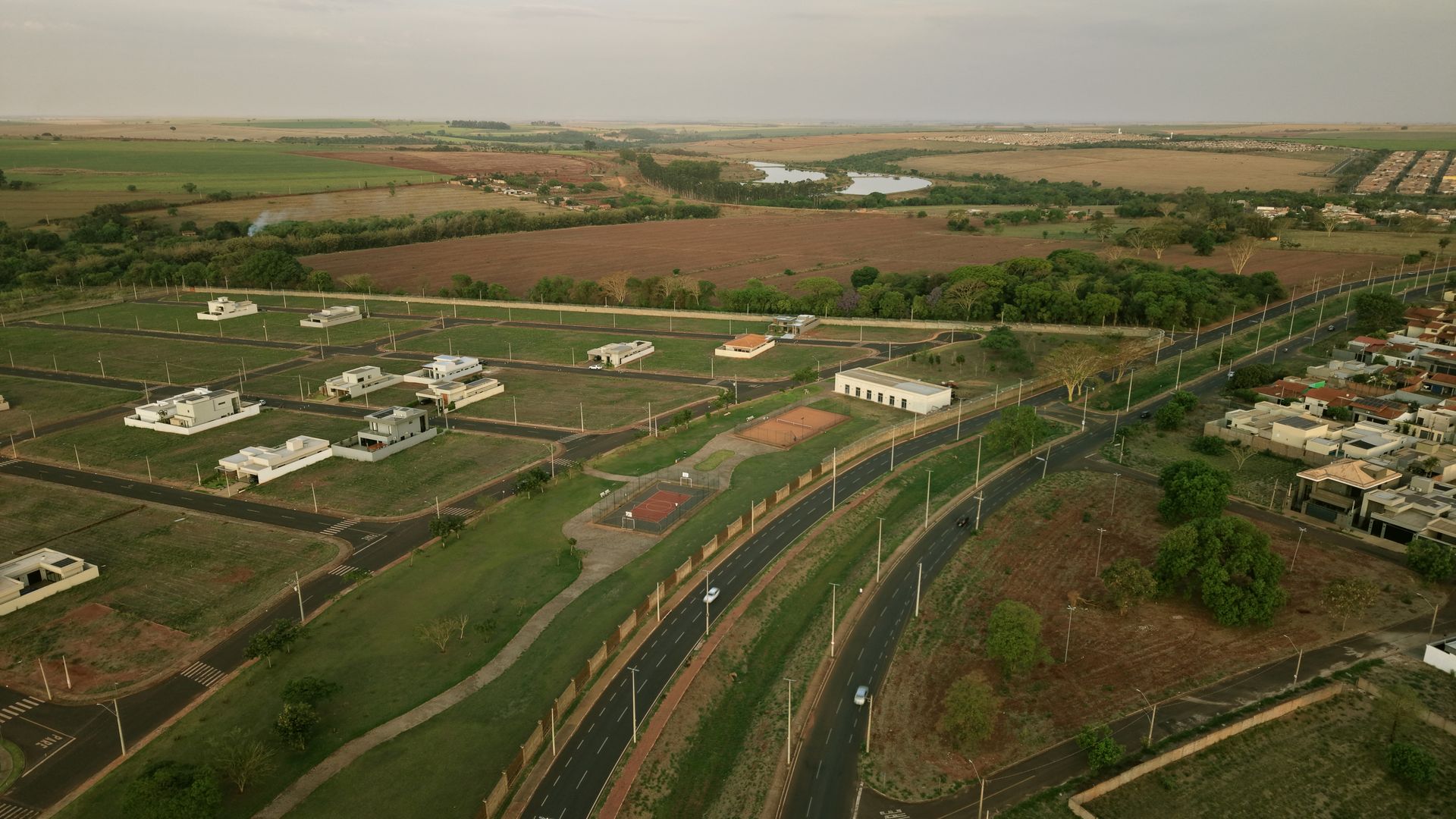 Vista aérea de uma área residencial próxima a uma rodovia, campos e um corpo d'água sob um céu nublado.