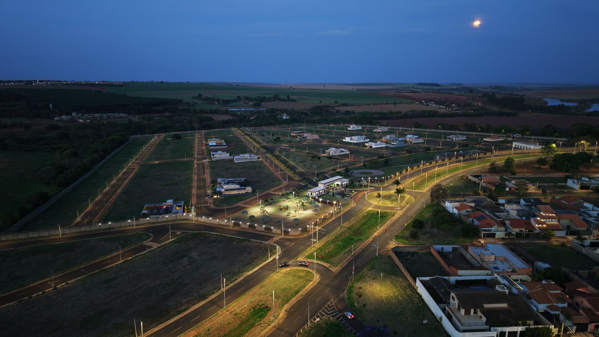 Vista aérea noturna de uma área residencial com ruas e casas iluminadas, sob um céu escuro com luar.