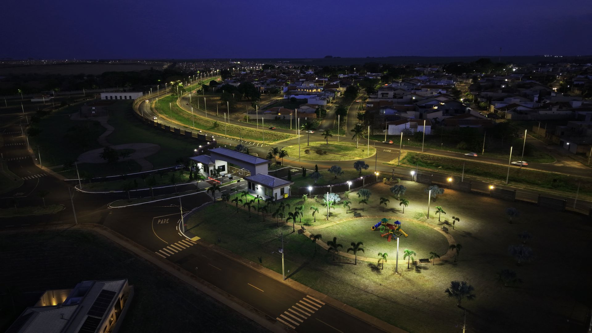 Vista aérea noturna de um parque com caminhos e edifícios iluminados, e uma área residencial ao fundo.