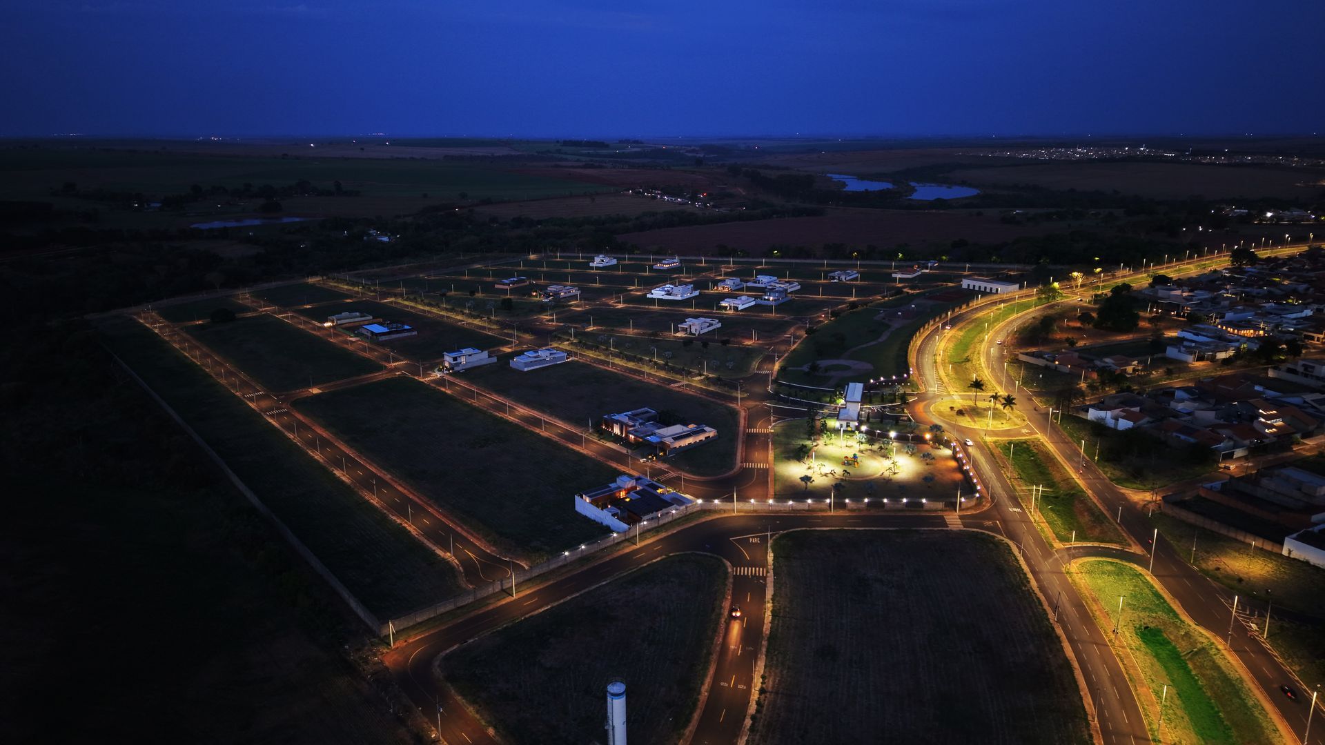 Vista aérea noturna de uma área residencial com ruas e casas iluminadas sob um céu azul escuro.