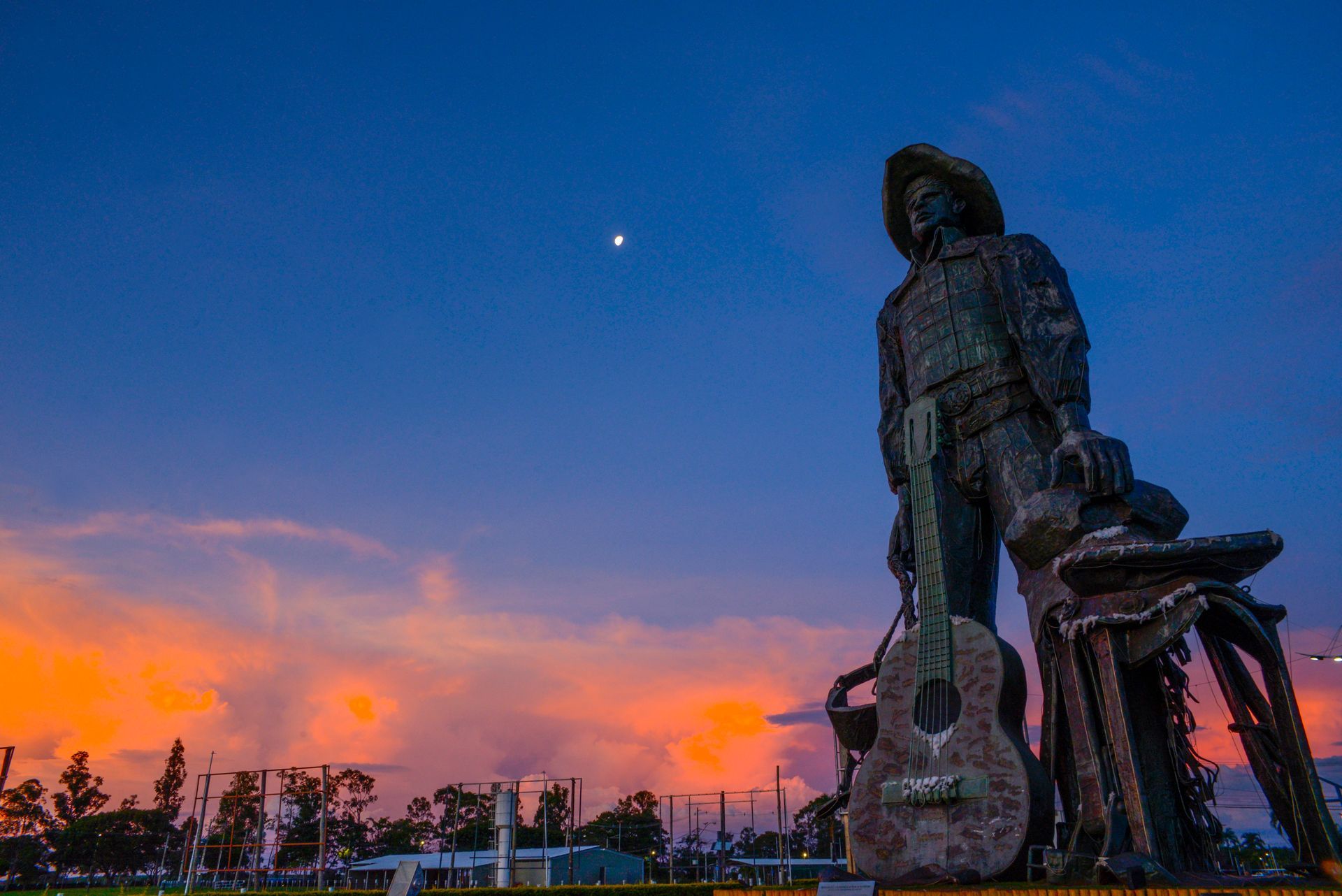 Estátua de um homem com guitarra e sela contra um céu ao pôr do sol.