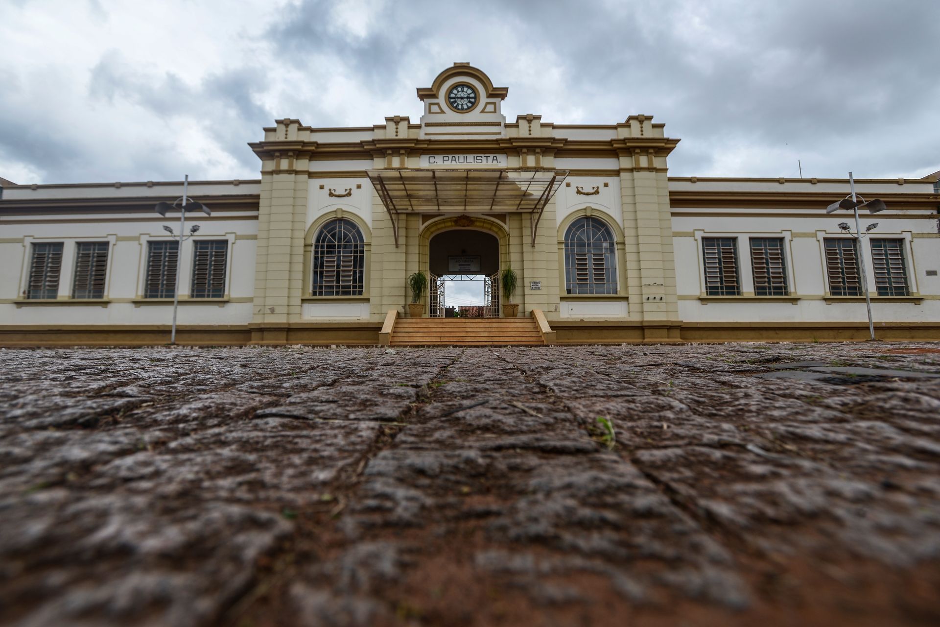 Vista exterior de um prédio antigo com colunas e relógio, sob um céu nublado. Tijolos marrons em primeiro plano.