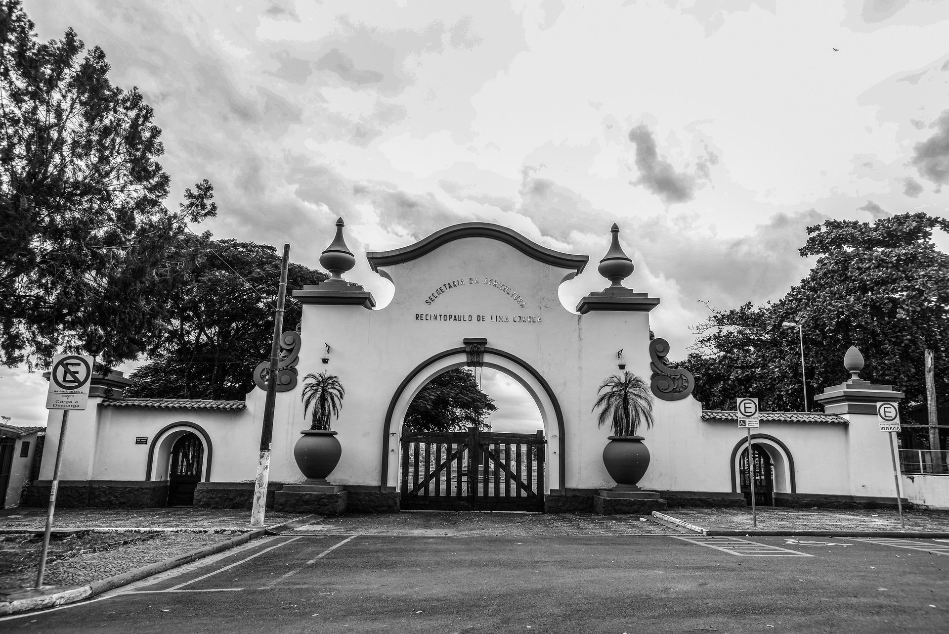 Portão branco ornamentado com arco e elementos decorativos, ladeado por árvores e uma estrada, em preto e branco.