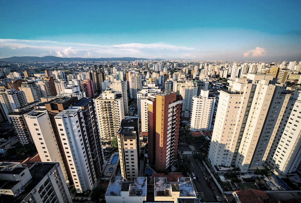 Vista panorâmica da cidade com inúmeros arranha-céus sob um céu azul; alguns edifícios são laranja e brancos.