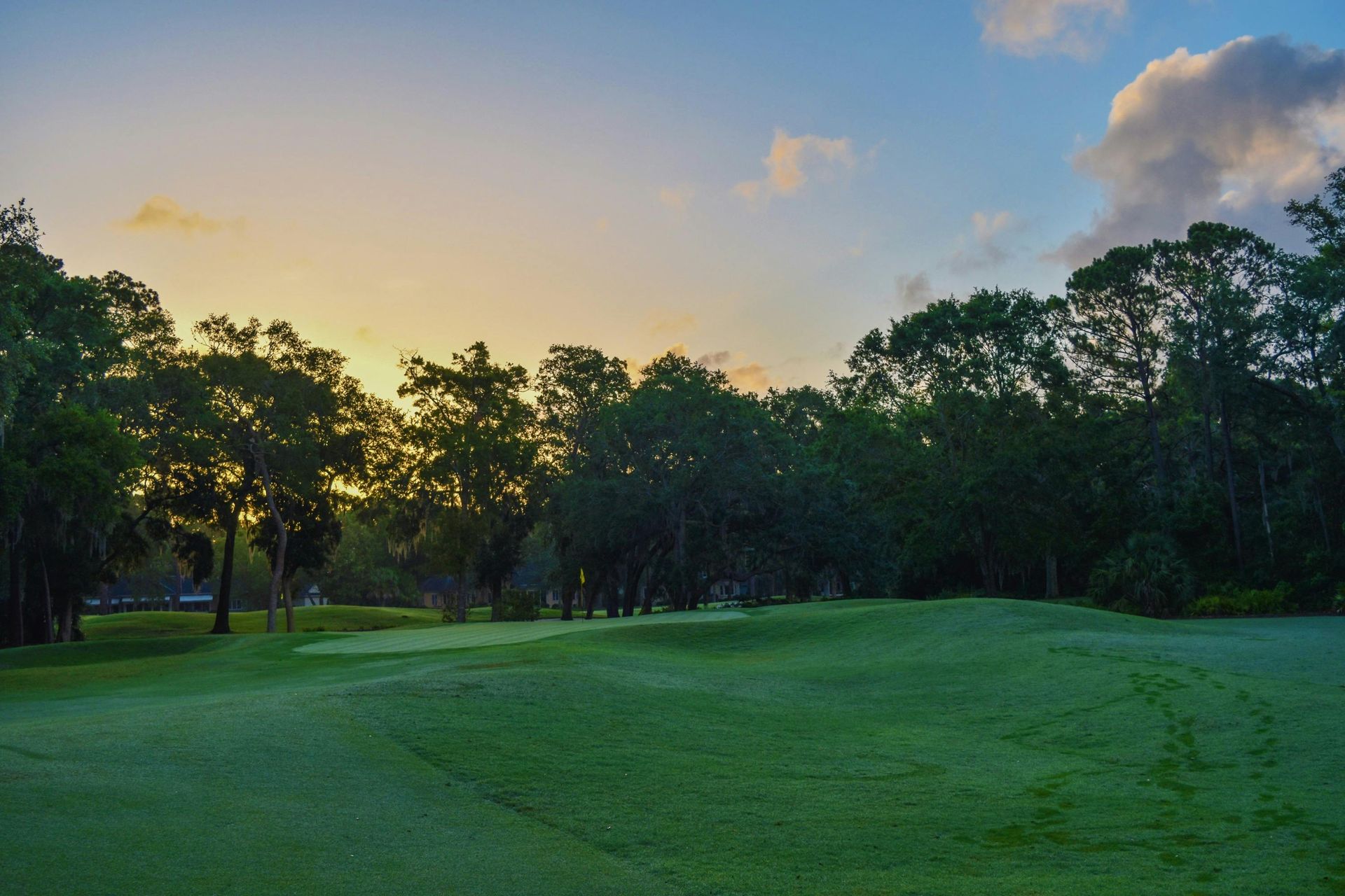 Campo de golfe ao amanhecer, com árvores e grama verde sob um céu colorido.