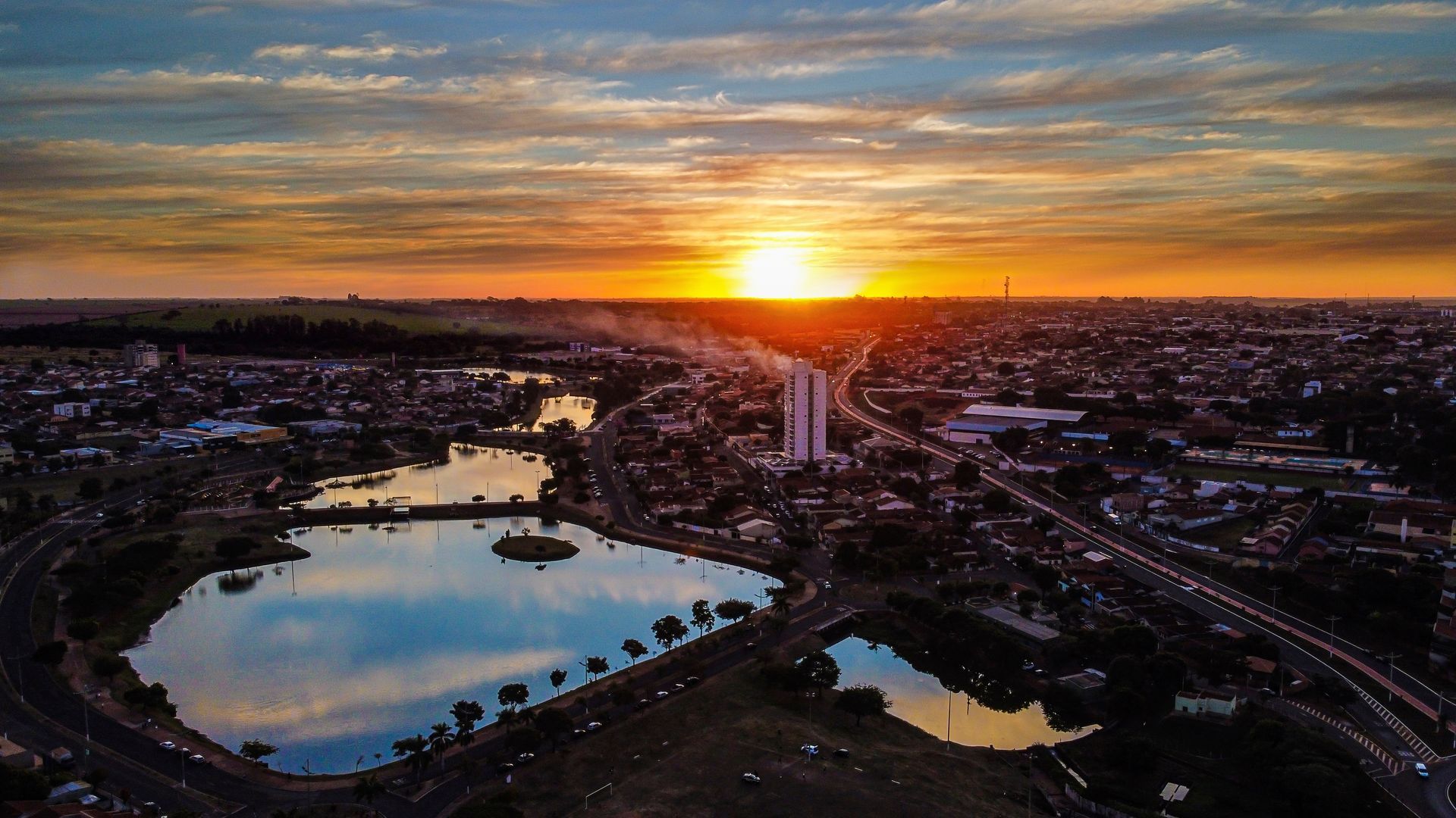 Paisagem urbana ao pôr do sol, com um lago refletindo o céu alaranjado e amarelo.