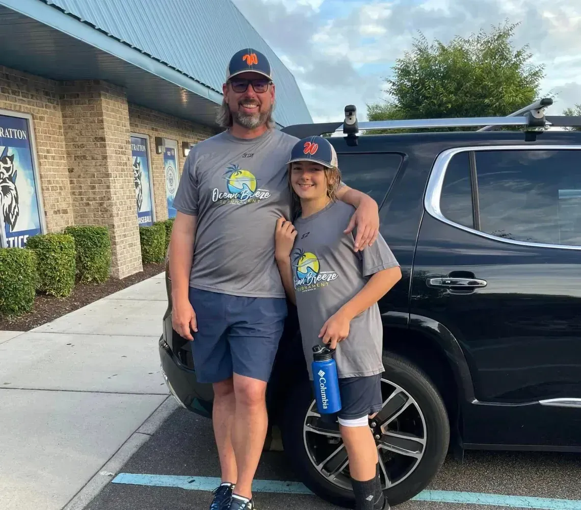 Two people in matching gray shirts and caps posing beside a black SUV outside a building.