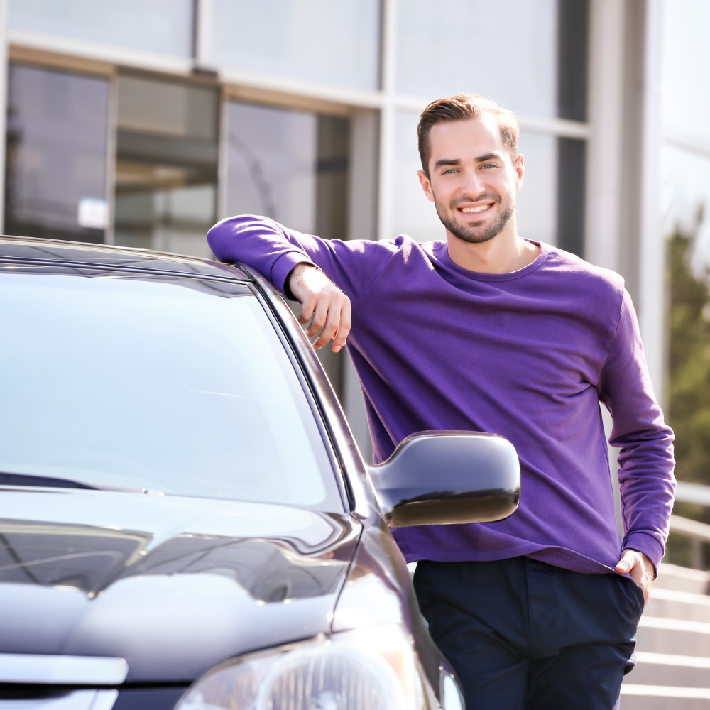 Man leaning on a black car, smiling outdoors in front of a building.
