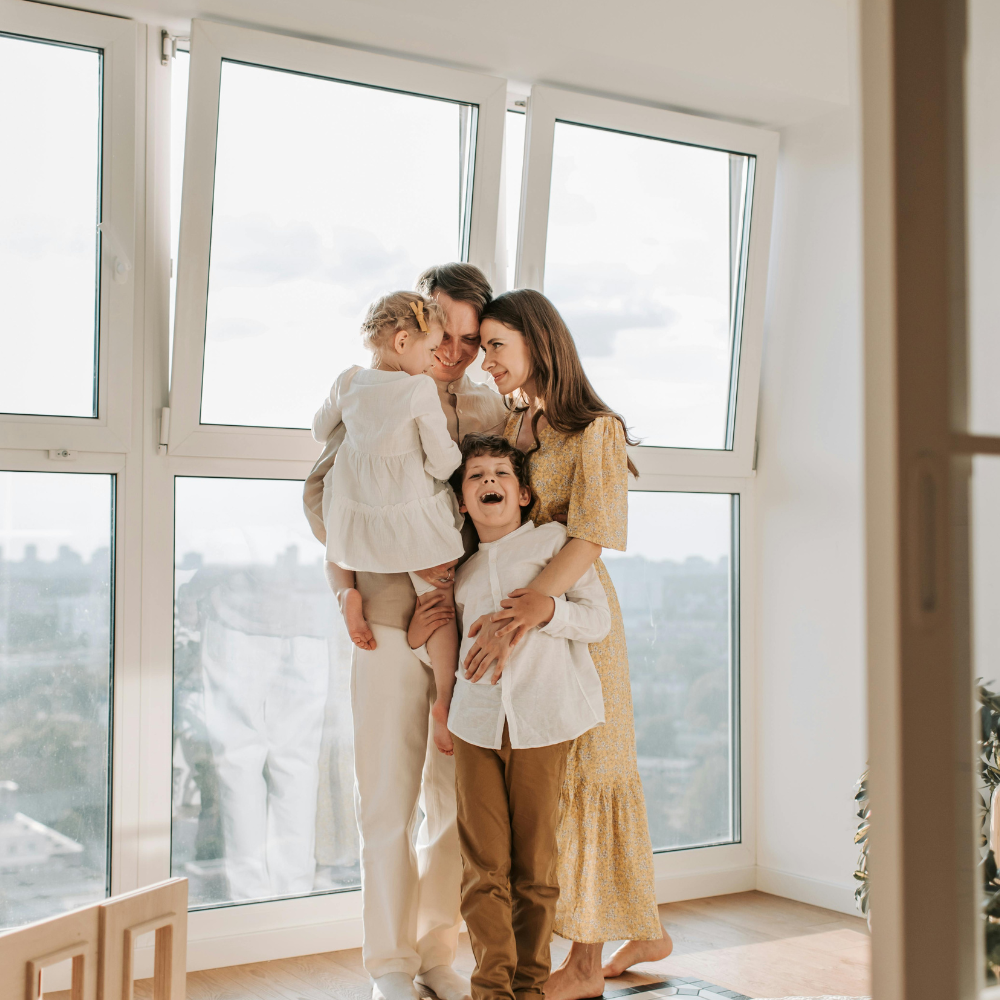 Family of four standing together in a bright room by large windows, embracing and smiling