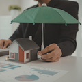 Hand holding a miniature house under a green umbrella on a desk with charts