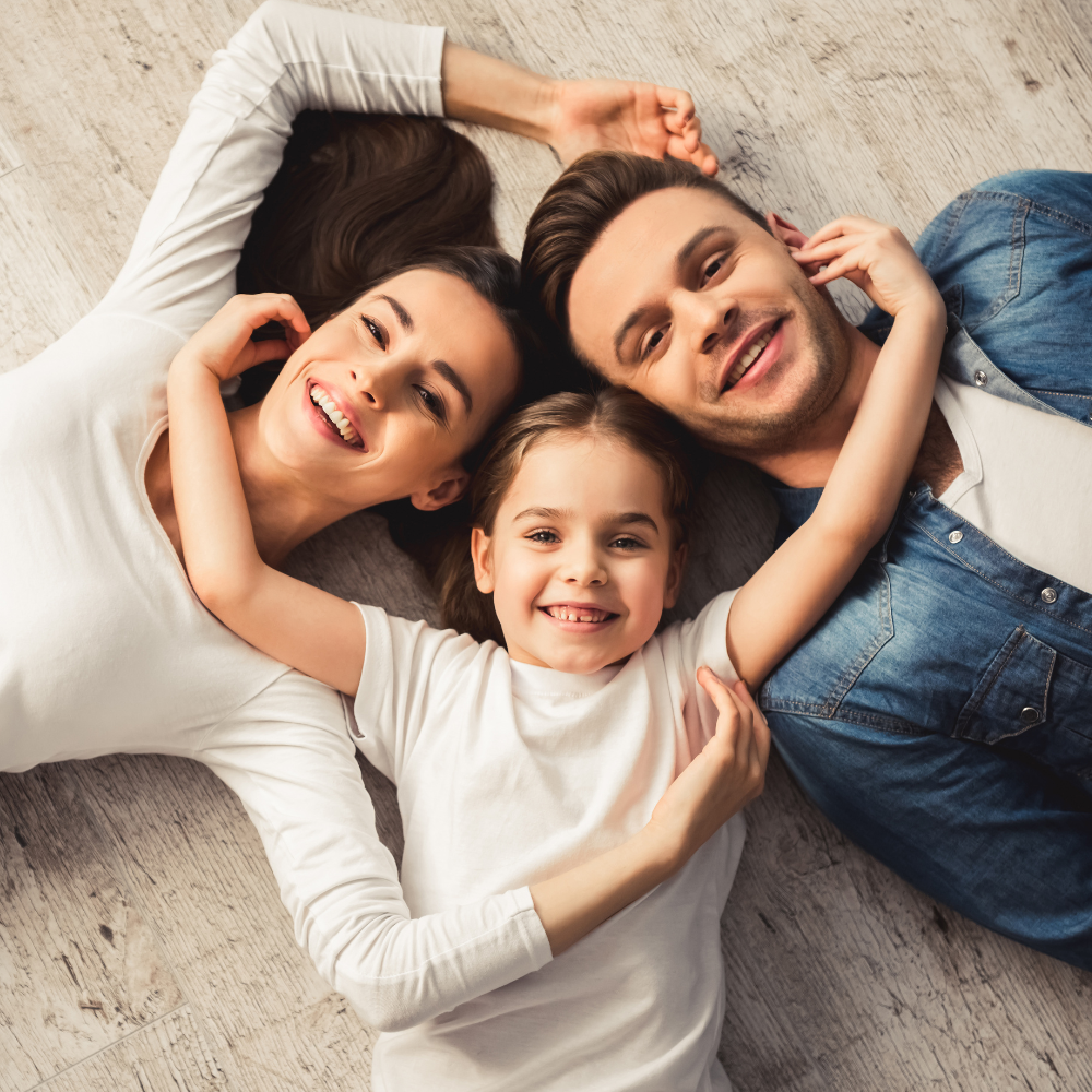 Three people lying on a rug, smiling and forming a heart shape with their arms
