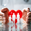 Hands holding a red heart behind a white family silhouette on a reflective surface